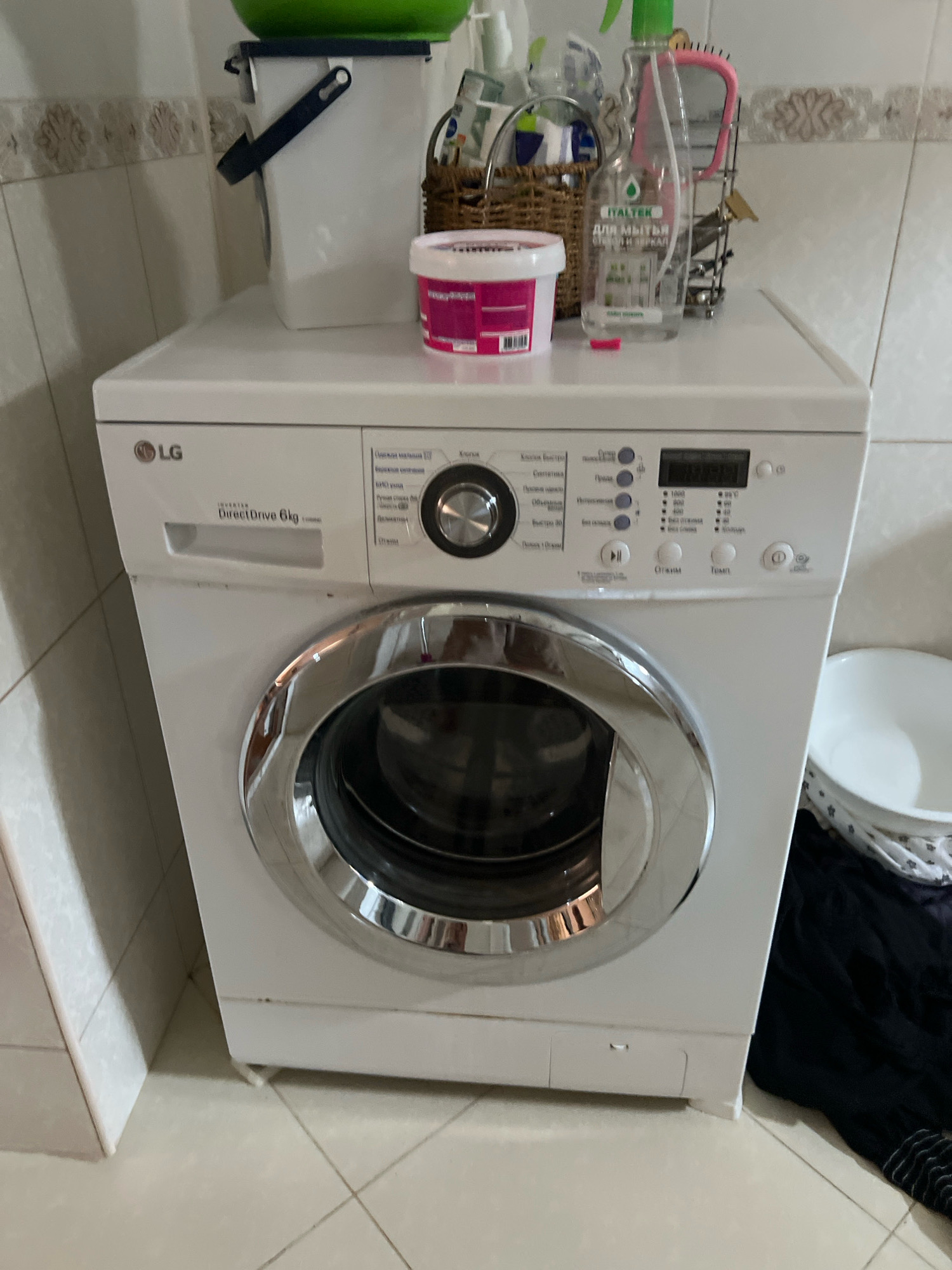 A front-loading LG washing machine is centrally located in a bathroom. The washing machine is white with a silver drum and control panel. Various cleaning supplies are arranged on top of the washing machine and on a nearby shelf. A wicker basket holds a pink-handled comb and a bottle of cleaning spray. Next to the washing machine, a white laundry basket contains dark-colored clothing. The bathroom features tiled walls and floor. The scene is static and captures a typical domestic setting.