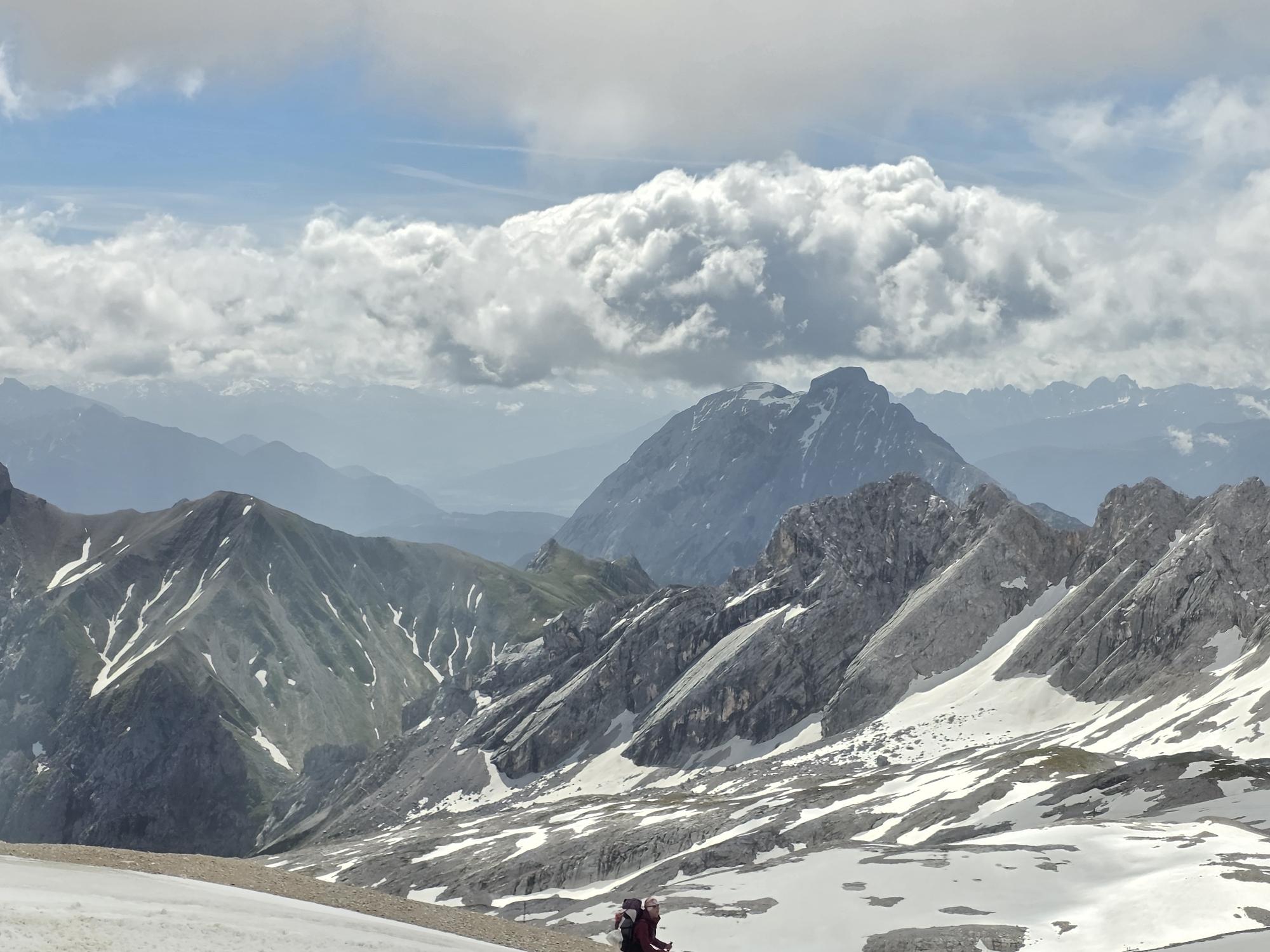 In the image, a panoramic view of the Zugspitze mountain range near Garmisch-Partenkirchen, Germany, is presented. The scene depicts rugged, snow-dusted mountain peaks under a partly cloudy sky. The foreground is a snow-covered slope with patches of rock and gravel. Two individuals, equipped with backpacks and hiking poles, are visible on this slope, seemingly engaged in a mountain trek. The middle ground showcases more imposing, rocky mountain formations interspersed with snowfields. In the background, successive layers of mountains fade into a hazy blue distance, with a prominent, distinct peak dominating the skyline. The sky is a mix of blue patches and large, cumulus clouds.