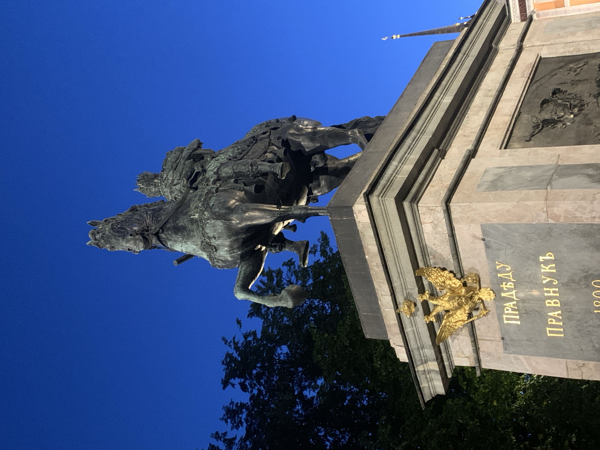 The image shows a bronze equestrian statue of Peter the Great, known as the Bronze Horseman, located on Senate Square in Saint Petersburg, Russia. The statue depicts Peter I on horseback, with the horse rearing on its hind legs. The monument is situated on a large granite pedestal, known as the Thunder Stone. The statue is illuminated, and the sky above is a deep blue, suggesting it is either dawn or dusk. In the foreground, there are inscriptions in gold lettering on the pedestal, and a golden double-headed eagle emblem is visible. Trees are visible to the left of the statue, and a spire of a building can be seen in the background to the right. The overall scene captures a significant historical monument against a twilight sky.