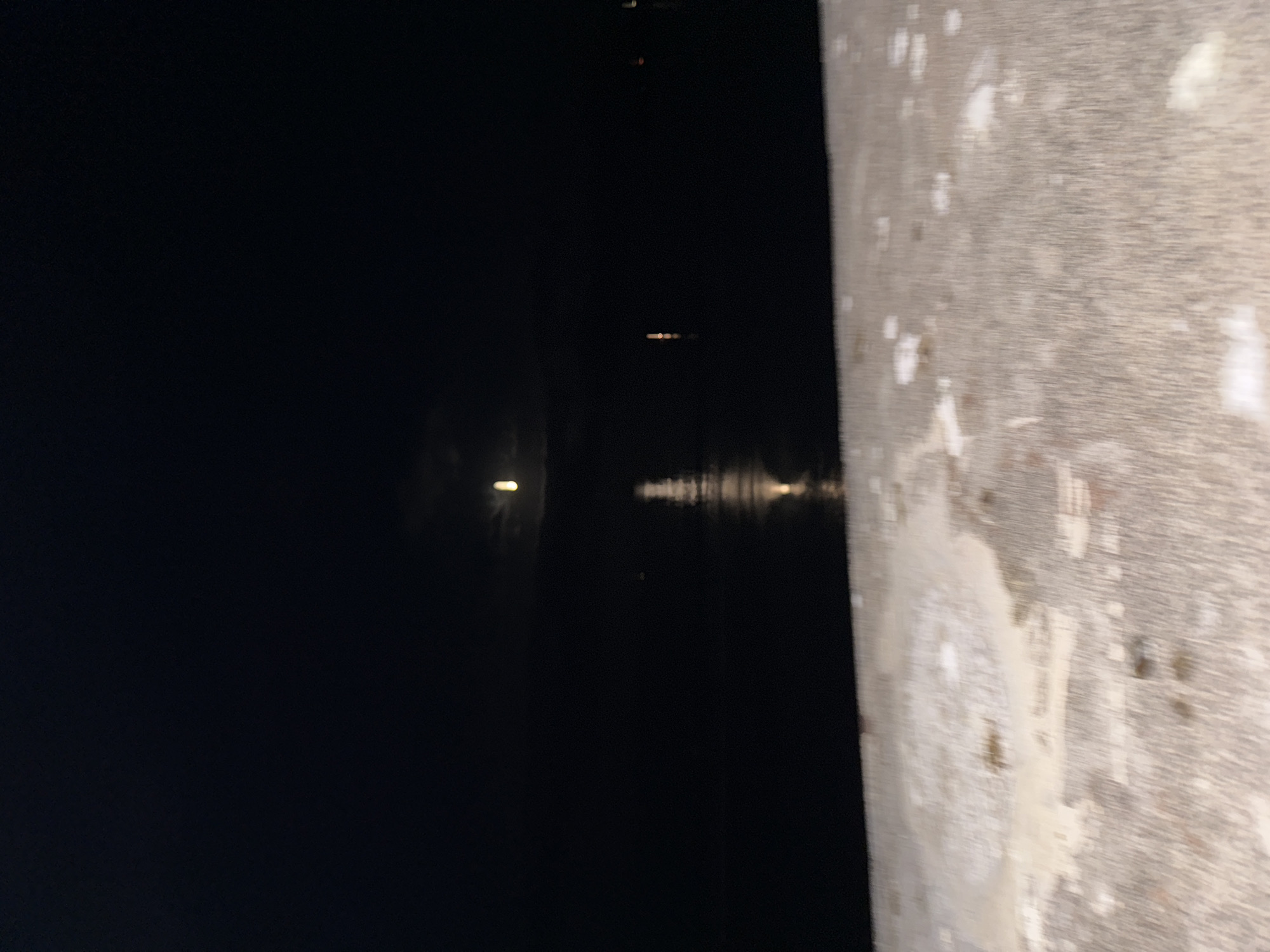 The image depicts a dark, nighttime scene with a body of water and a faint reflection of the moon. In the foreground, a textured surface, possibly a railing or wall, is visible with patches of lighter material that could be frost or snow. The water appears calm, with a single bright reflection of what might be a light source or the moon itself. The sky is a deep black, indicating nighttime. The location is identified as Tramore, Ireland, suggesting a coastal or waterfront setting. There are no people or discernible actions taking place in the image. The overall impression is one of a quiet, cold, and dark night.