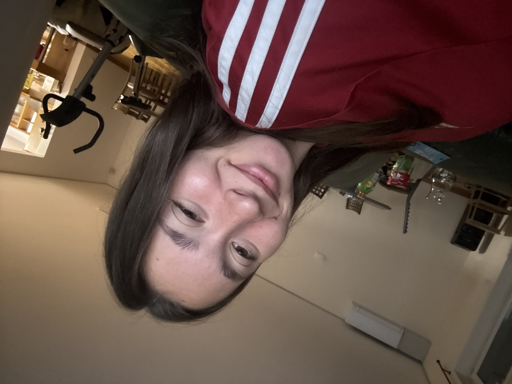 A young woman with long, dark hair is taking a selfie in what appears to be a living room or common area. She is wearing a red shirt with white stripes on the shoulder and sleeve. The room is lit by artificial light, creating warm tones. In the background, an exercise bike is visible, along with a dining table and chairs. Other objects like a shelf with a speaker, a glass of water, and some packaging are also present. The overall impression is a casual indoor setting.
