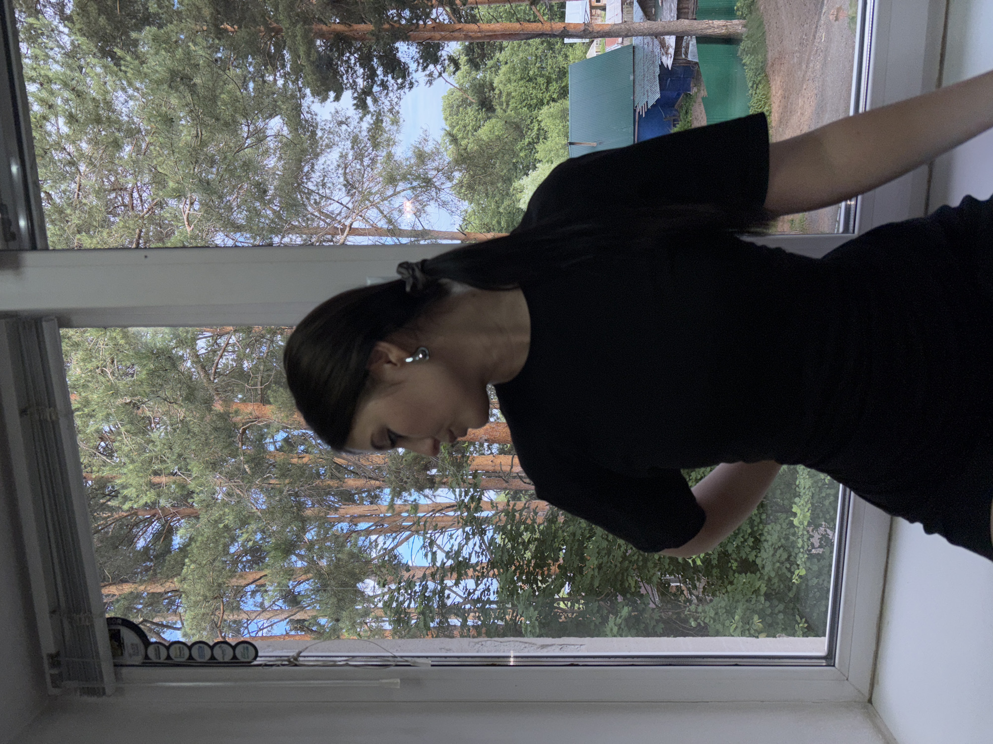 A young woman with dark hair tied in a ponytail, wearing a black t-shirt and shorts, is standing with her back to the camera, looking out a window. She is positioned on a windowsill inside a building. Outside the window, a dense forest of pine trees can be seen, with a few houses and a green fence in the background. The sky is partly cloudy. The scene is likely located in Chaikovsky, Russia, given the characteristic pine forests and dachas often found in the region.