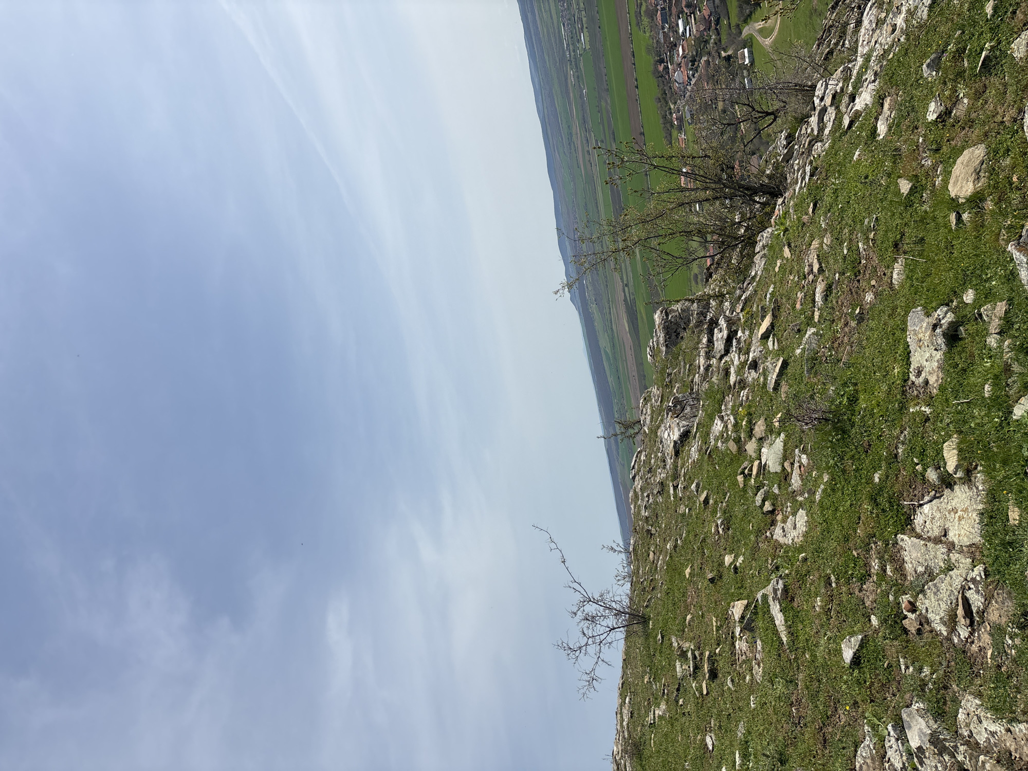 The image presents a high-angle view from a rocky, grassy hillside. The terrain is interspersed with light-colored rocks and green vegetation. In the distance, a landscape of green fields and a settlement with houses and roads is visible. The sky takes up a significant portion of the frame, showing a gradient from a light blue to a pale, cloudy white. Bare trees or bushes cling to the hillside. The location is Goritsa, Bulgaria. No people are visible.