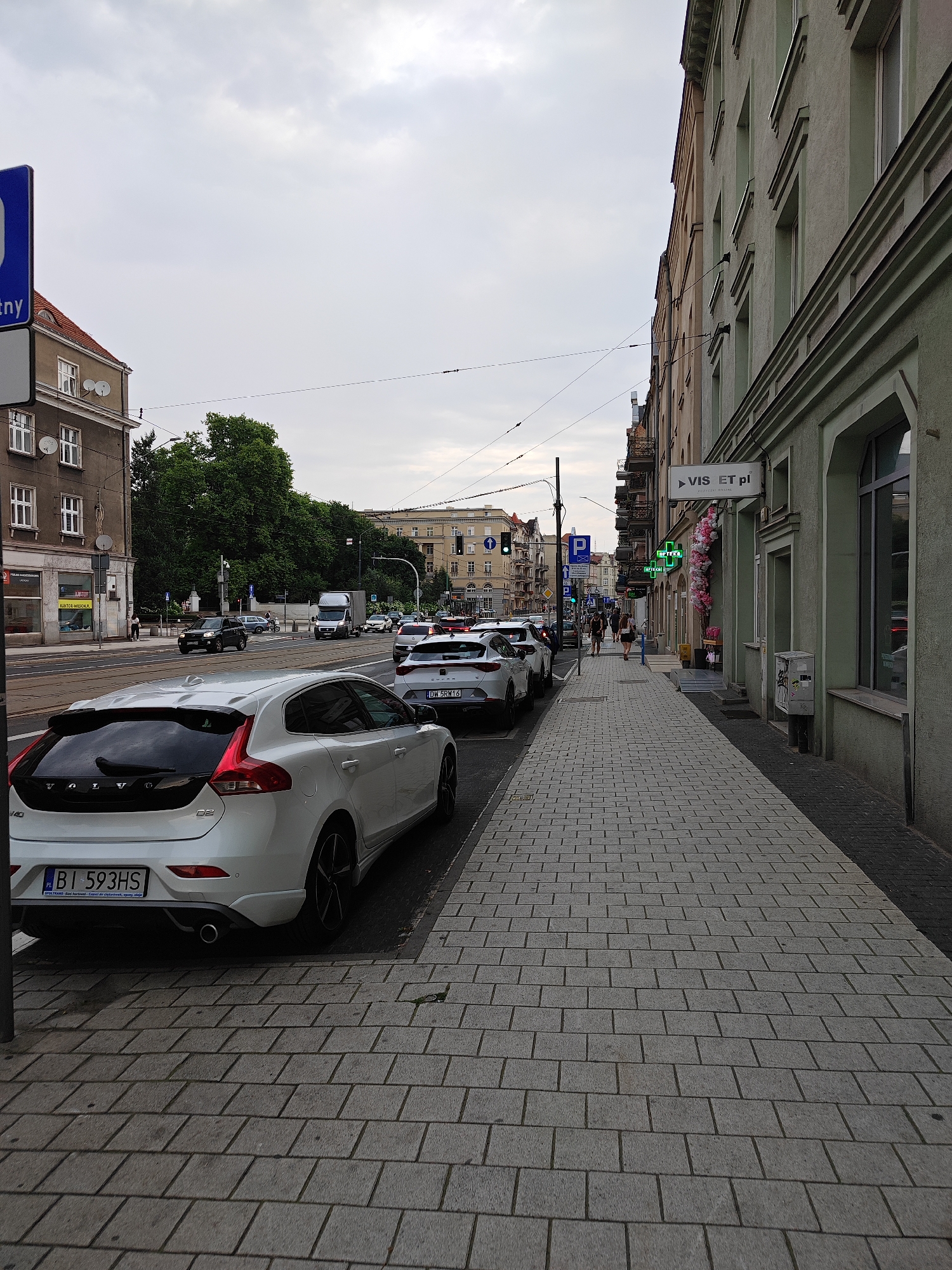 The image shows a street scene in Poznan, Poland, with a focus on a sidewalk and parked cars. On the right side of the image, a light green building with multiple windows lines the sidewalk, which is paved with grey rectangular stones. A sign reading "VIS ET pl" is visible on the building. Further down the street, a green cross sign indicates a pharmacy. On the left side of the image, a white Volvo hatchback is parked on the street, facing away from the camera. The car's license plate reads "BI 593HS". Across the street, there are other vehicles, including cars and a truck, and some buildings, including a red-roofed building. Trees are visible in the background. The sky is overcast. A person is walking on the sidewalk in the distance.