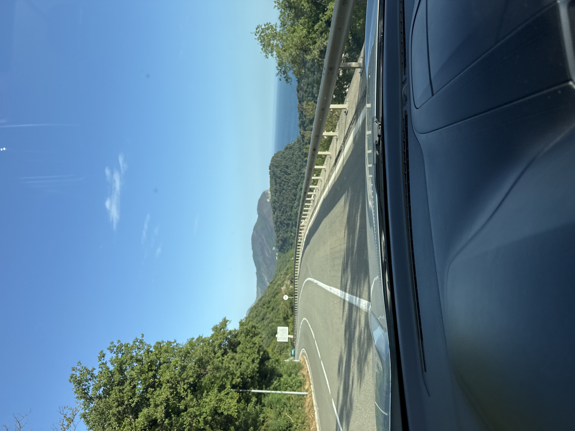 The photograph, taken from inside a vehicle, depicts a curving road with a guardrail. Green trees and vegetation border the road. In the distance, there is a view of a coastline and the sea. The sky is clear and blue. This road is located in the city of Stupovi, Montenegro.
