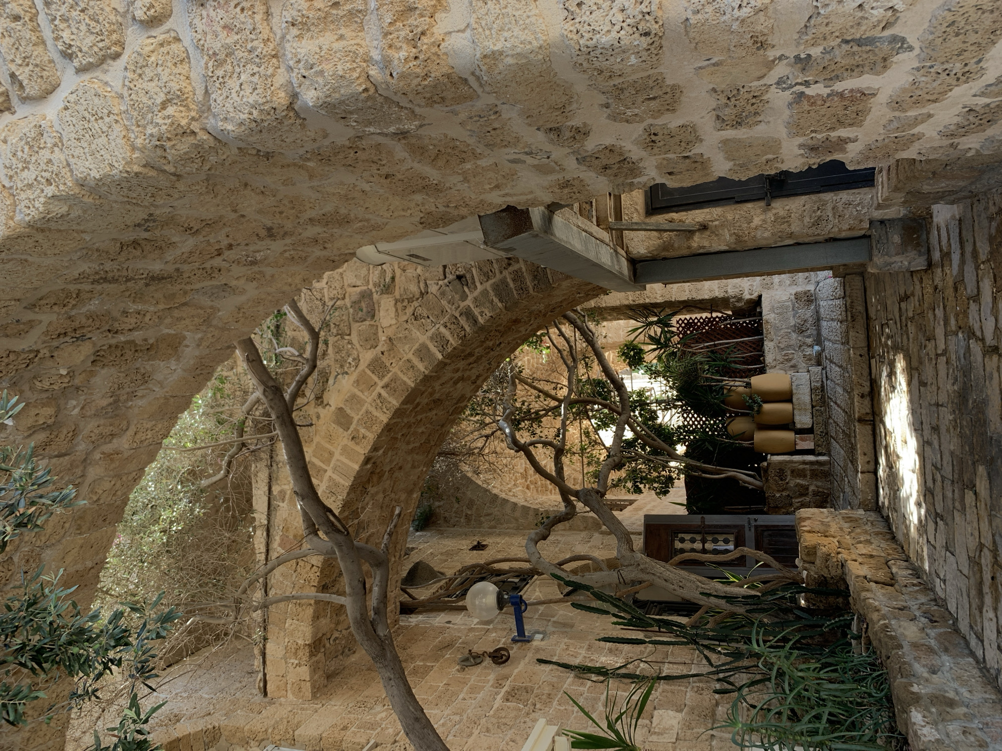 The image depicts an arched stone passageway in the ancient city of Jaffa, which is now part of Tel-Aviv, Israel. The passageway is constructed from rough-hewn, light-colored stones, and appears to be part of an old building. Sunlight streams through the archways, casting dappled shadows on the stone-paved ground.
In the foreground, to the left, a stone wall lines the walkway, topped with a variety of plants, including tall, spiky green plants and a larger tree with gnarled branches. A vintage-style light fixture with a spherical globe is mounted on the stone wall. A wooden door is visible further down the passageway on the left.
To the right, the stone wall of the passageway continues, with another archway leading further into the distance. A dark metal door is set into this wall. In the background, beyond the archways, more greenery and stone structures are visible, suggesting a dense urban environment with historical architecture. The overall impression is one of a secluded and historic alleyway in a Mediterranean setting. There are no people visible in the image.