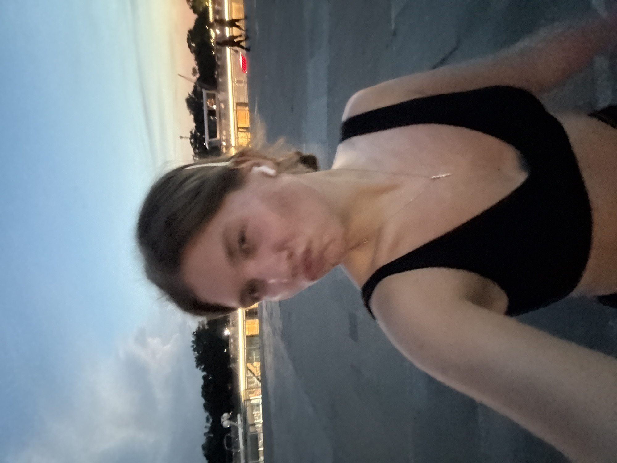 A young woman is taking a selfie outdoors at dusk. She is wearing a black sports bra and a silver necklace. Her hair is pulled back, and she is wearing white wireless earbuds. She is making a pouting face. In the background, there is a body of water with a boat docked along its edge, and several people are walking on a promenade. The sky is a twilight blue with some clouds. The location appears to be the Sava Promenade in Belgrade, Serbia, with the Sava River and the Gardoš Tower visible in the distance.