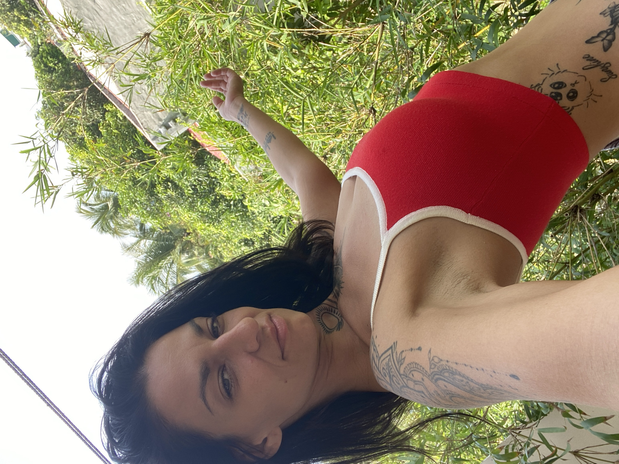 A woman with dark hair and multiple tattoos is posing for a selfie outdoors. She is wearing a red tank top with white trim. She is leaning to her left, with her right arm outstretched and her hand open, reaching towards a cluster of bamboo plants. Behind her, there is a building with a gray roof and a wall covered in white plaster. The background is filled with lush green vegetation, including palm trees, suggesting a tropical or subtropical climate. The sky is bright and overcast. The scene appears to be a residential area with dense foliage, possibly a backyard or garden. There is no indication of the specific location within the city of Mũi Né, Vietnam, beyond the general outdoor setting.