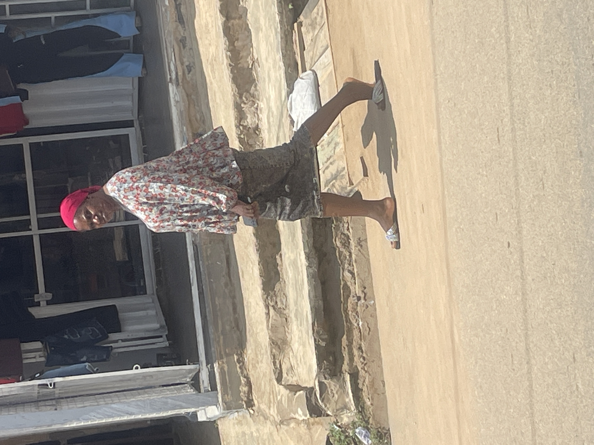 A woman in a floral print top and a patterned skirt walks on a paved street in Abuja, Nigeria. She is wearing a pink headscarf and sandals. Behind her are steps leading up to a building with a large glass window. A white bag is visible on the ground next to the steps. The area appears to be a marketplace or a commercial street.