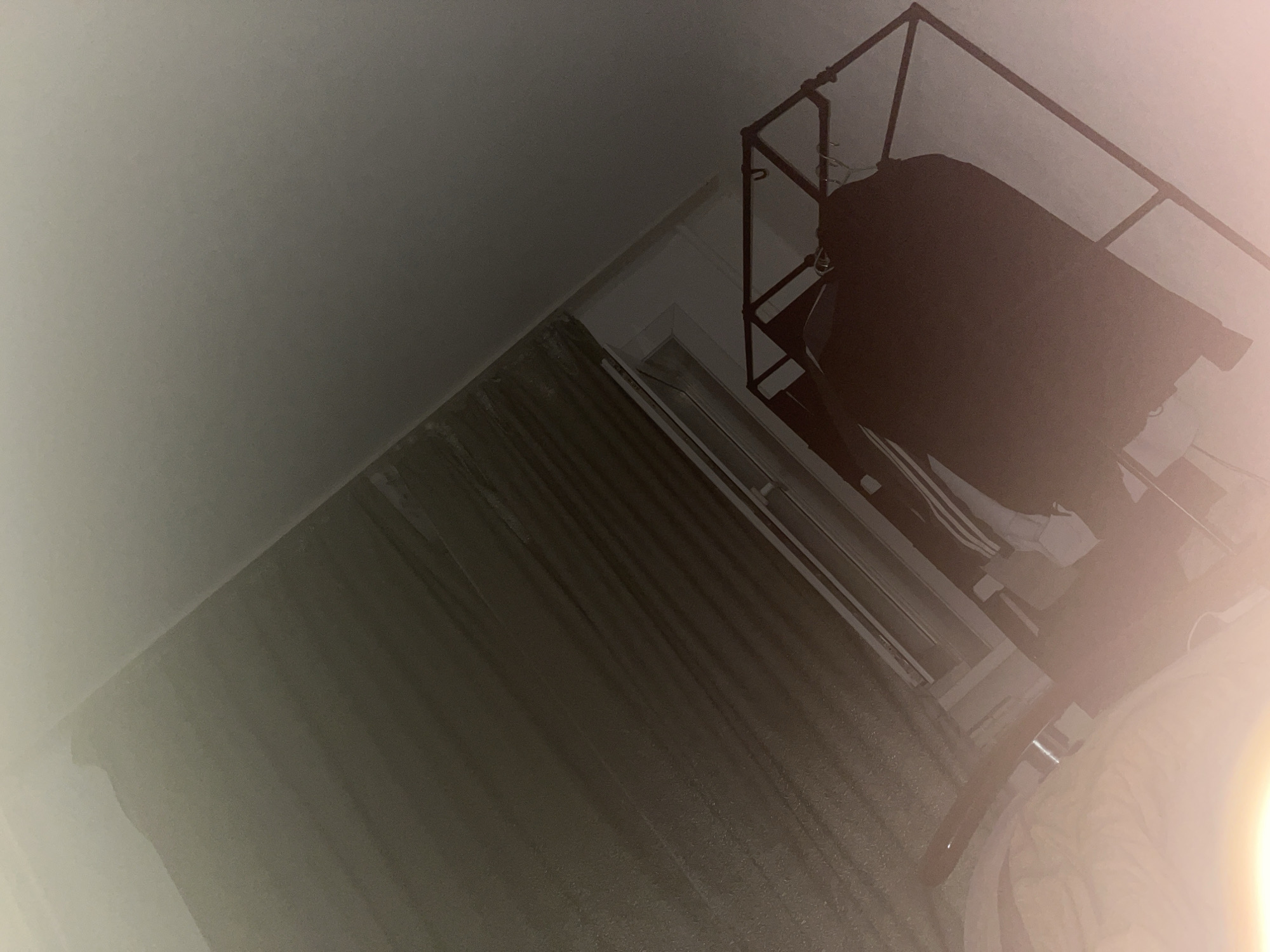 The image depicts an indoor scene, possibly a bedroom, with a low-angle perspective. The dominant elements include a gray ceiling, a diagonally oriented wall with curtains or blinds, and a black metal rack holding a dark garment. A bright light source, possibly a lens flare or flash, obscures parts of the right side of the image. The environment appears to be dimly lit, with a muted color palette. The location in Neryungri, Russia, is not visually ascertainable from the provided image.