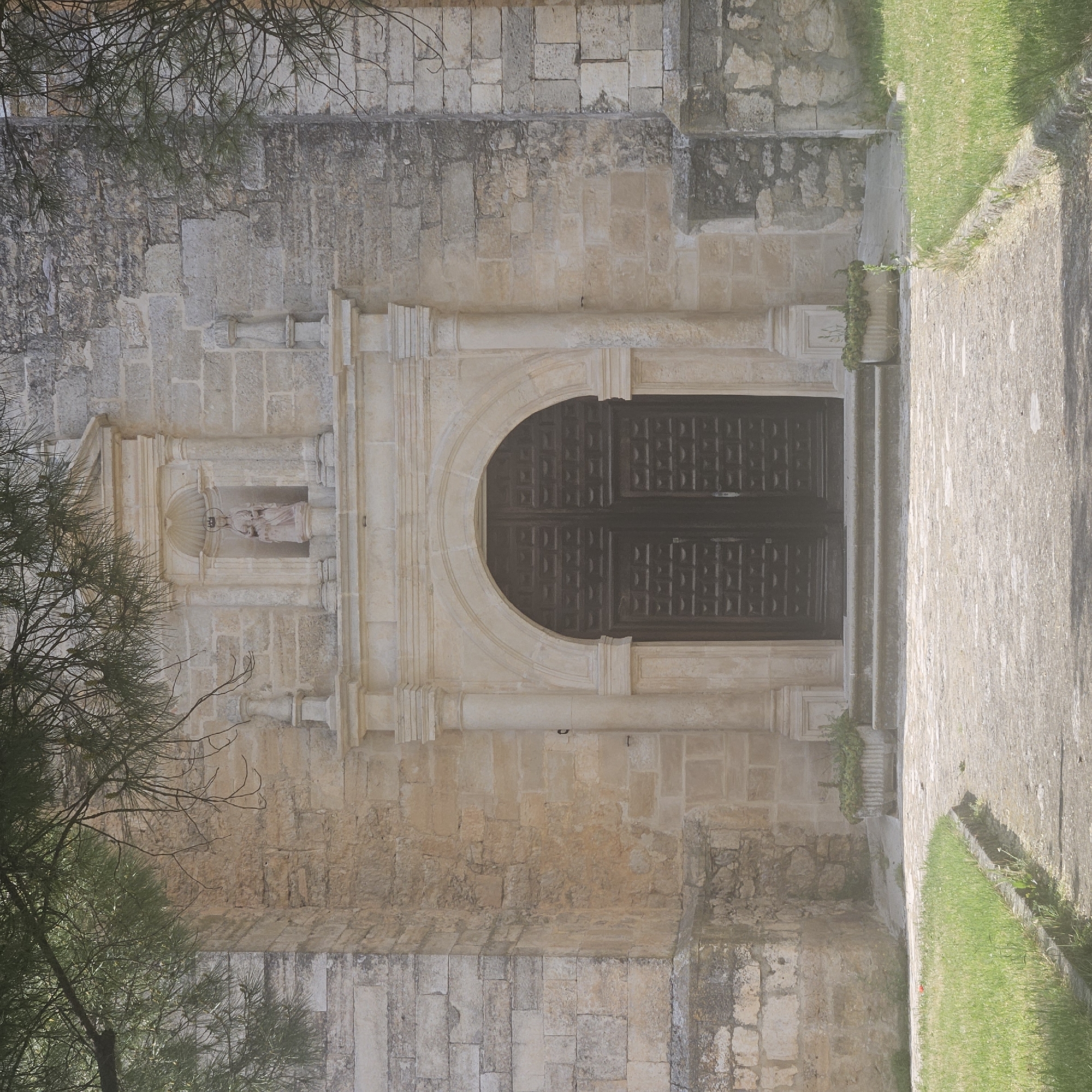 The image presents the exterior of the Church of San Pedro Apóstol in Algora, Spain. The facade, constructed from light-colored stone, features an arched doorway with decorative molding. Above the doorway, a niche contains a statue of the Virgin Mary. The building's exterior displays a weathered texture. A stone pathway runs alongside the church, bordered by grass. A tree partially obscures the left side of the frame.