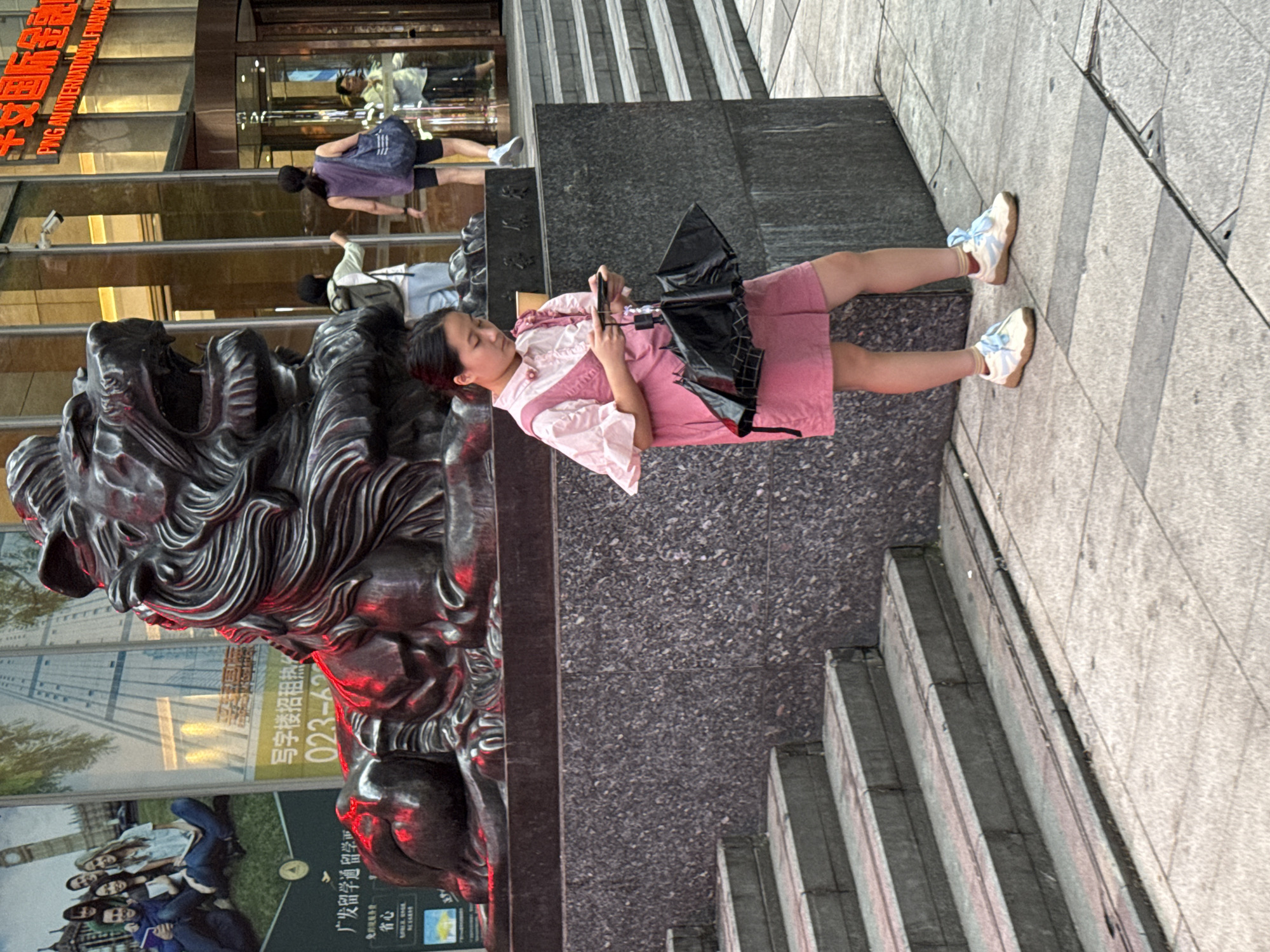 A young East Asian girl, appearing to be in her early teens, stands on a sidewalk in front of a large, dark stone lion sculpture. She wears a pink short-sleeved dress over a white ruffled collar shirt and white sneakers with light blue laces and trim. She is holding a smartphone in her right hand and a black umbrella in her left, which is partially open. The lion sculpture is illuminated by red lights from below. Behind the girl and the sculpture is a building with large glass windows, some reflecting lights. To the left of the sculpture, a large advertisement is partially visible, featuring images of young people. A set of concrete stairs is positioned to the left of the frame. In the background, several people are visible entering and exiting the building. The scene appears to be in a commercial area of Yuzhong District, China, possibly in front of a financial institution, indicated by signage that includes "PING AN INTERNATIONAL FINANCIAL".