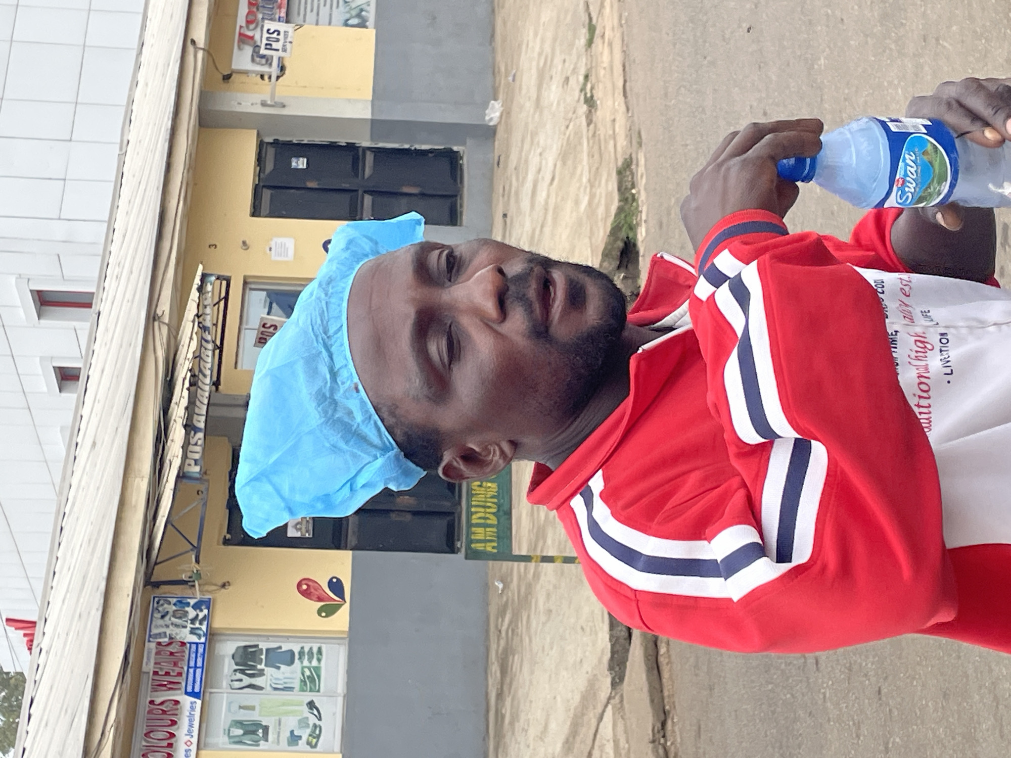 A man wearing a light blue surgical cap and a red and white track jacket drinks from a clear plastic water bottle labeled "Swan." He stands on a sidewalk in front of a building. The building has a sign indicating "COLOURS WEARS" that sells clothes and jewelry. Another sign above the building reads "POS AVAILABLE." Green signage with the words "AM DUNG" is also visible. The location is likely in the city of Jos, Nigeria, based on the local business name and visual cues of the environment.
