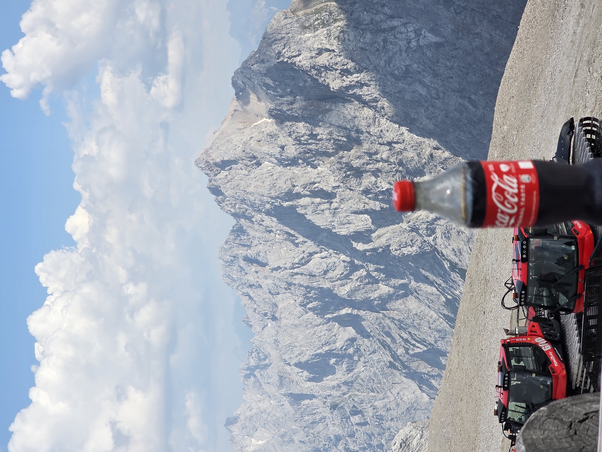 The image shows a close-up of a partially filled Coca-Cola bottle in the foreground, with a mountainous landscape in the background. The bottle has a red cap and a white and red Coca-Cola label. The mountain range is rugged and rocky, with patches of snow visible. Two red snow grooming vehicles are parked on a gravelly slope to the right of the bottle. The sky is blue with scattered white clouds. The scene appears to be set in a mountainous region, likely during daytime given the bright lighting. The orientation of the image is sideways. Text visible on the Coca-Cola bottle reads "Coca-Cola" and "Original Taste." There is also text on the snow groomers, including "600" and "Zugspitzbahn". The location context suggests this could be the Zugspitze, Germany's highest peak, near Garmisch-Partenkirchen.