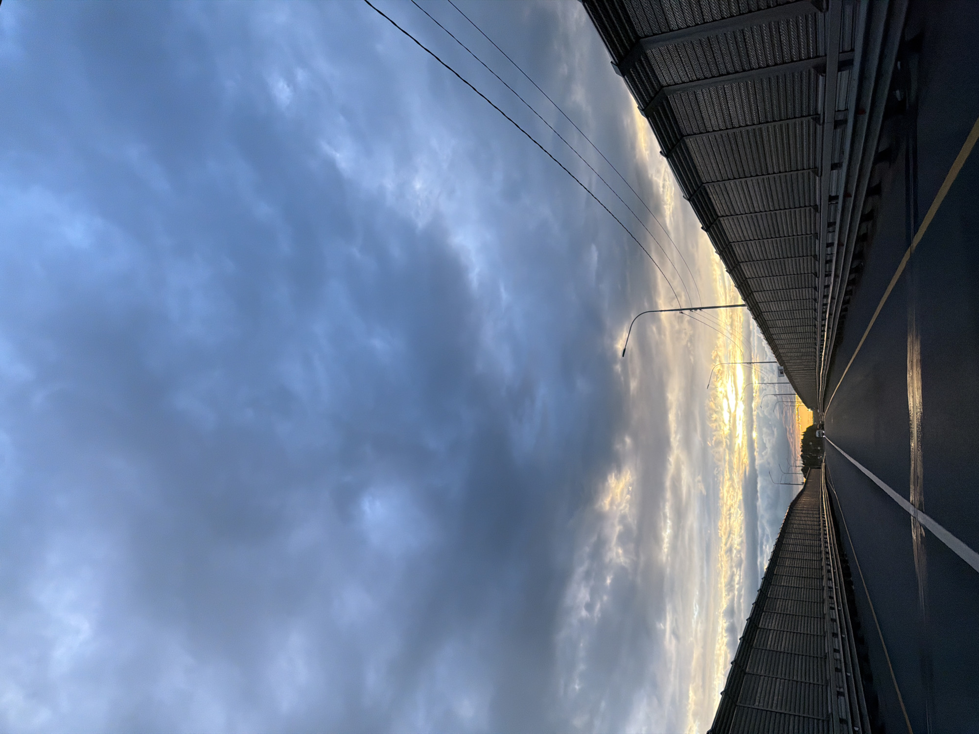 The image shows a view along a road or highway, presumably a bridge, with a sky filled with clouds above. The road surface appears dark, possibly wet, and features white and yellow lane markings. On either side of the road are structures, possibly sound barriers or railings. Street lamps are visible along the side. The sky contains a mix of blue and gray clouds, with areas of brighter, possibly sunset or sunrise colors, appearing through the clouds. Power lines are present above the road. The location and city are unknown.