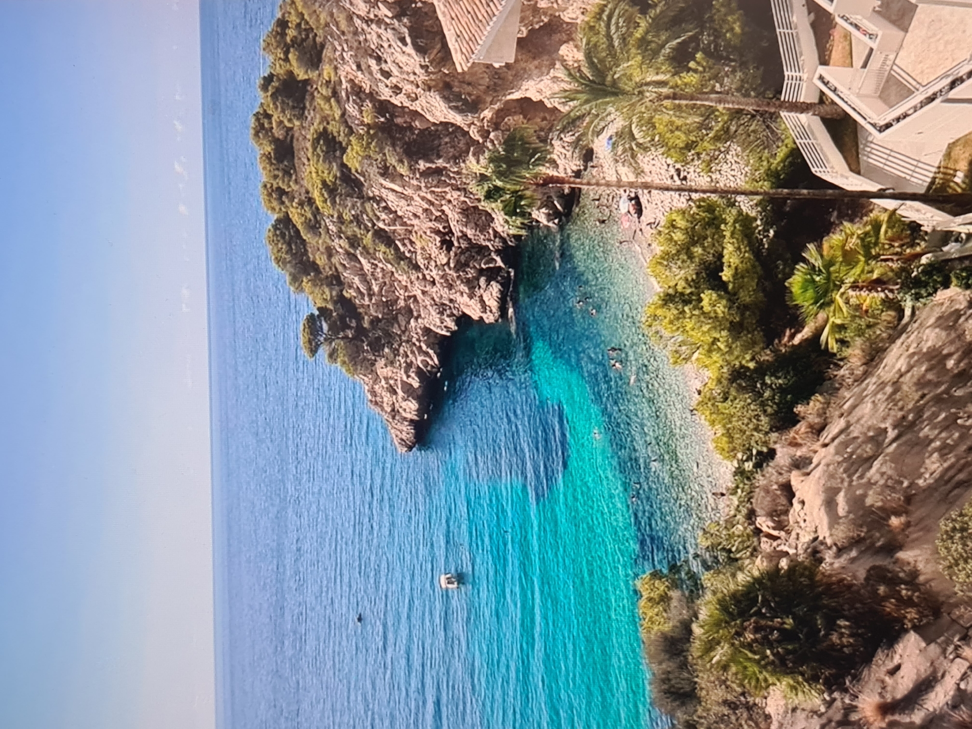 An aerial or elevated view captures a picturesque coastal cove on a bright, sunny day. The scene features clear turquoise waters, rugged cliffs, lush green vegetation, and architectural elements.
The sea dominates the left side of the image, transitioning from a deeper blue on the horizon to a vibrant, transparent turquoise closer to the shore. A small, light-colored boat is anchored in the deeper blue water. The cove itself cradles a light-colored beach, which appears to be a mix of sand and pebbles. Numerous small figures are visible in the water, engaged in swimming or wading, particularly in the shallower, clearer sections. A few people are also seen relaxing on the beach.
The cove is bordered by steep, rocky cliffs covered in dense green Mediterranean vegetation, including various trees and bushes. On the right side, modern white buildings with multiple levels and balconies are built into the cliffside, suggesting a hotel or residential complex. These buildings are surrounded by abundant greenery, featuring several prominent palm trees. Another structure with a reddish-brown tiled roof is visible on the upper part of the left-hand cliff.
The sky is a clear, bright blue with no visible clouds, indicating pleasant weather. The overall atmosphere is serene and inviting, characteristic of a vacation destination. No text is visible in the image.