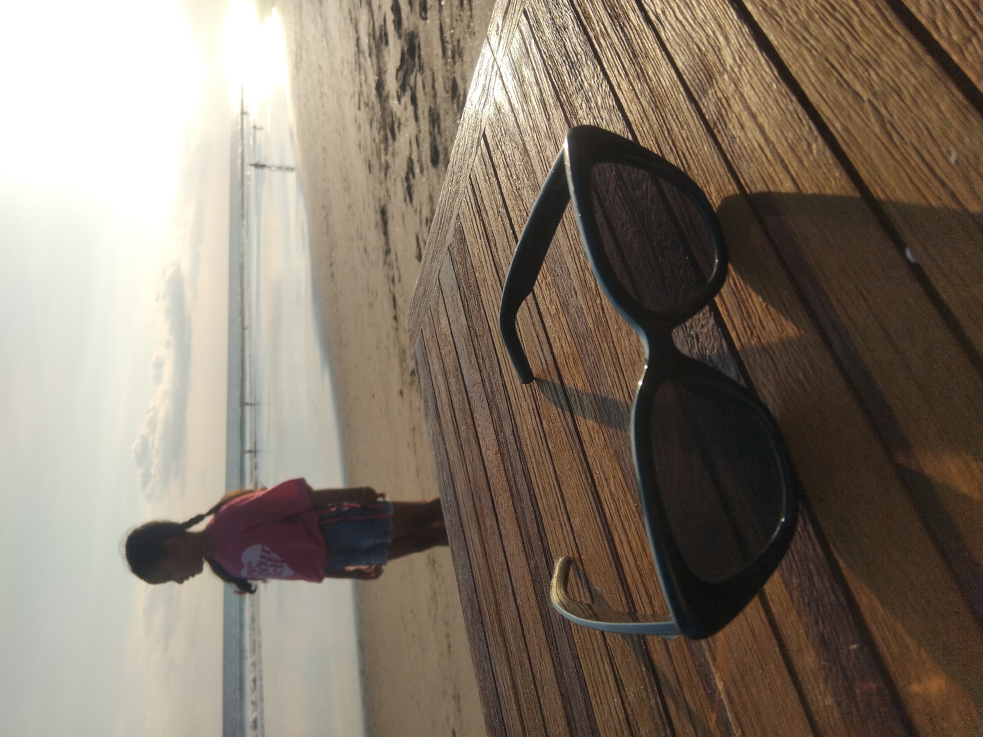 This image captures a tranquil beach scene, taken from a low perspective. In the foreground, a pair of black sunglasses with dark, reflective lenses is positioned on a slatted wooden surface, which exhibits a rich, warm brown tone. The strong, low sunlight creates distinct shadows from the sunglasses and highlights the grain of the wood.
Beyond the wooden surface, a sandy beach extends into the mid-ground, where a young girl stands. She has dark braided hair and is wearing a pink t-shirt with an indistinct white graphic, possibly a heart design, and light-colored denim shorts. She is silhouetted against the bright sky, facing away from the viewer and appearing to look towards the ocean.
The ocean meets the shore in the mid-ground, stretching towards a hazy horizon. The sky is bright with scattered clouds, indicating either early morning or late afternoon. The intense sunlight in the top left corner suggests a powerful sun, casting a soft, warm glow across the entire scene. Faint structures, possibly a distant pier or markers, are visible on the horizon line over the water. The overall atmosphere is peaceful and leisurely, characteristic of a beach setting in North Kuta, Indonesia.