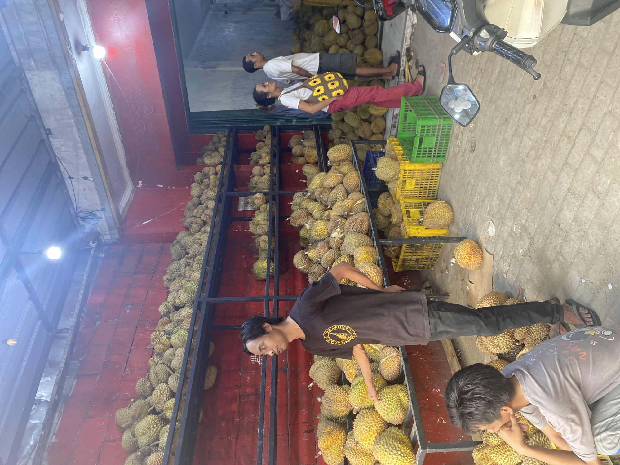 The image depicts an outdoor durian fruit stand in Depok, Indonesia. The stand is stocked with a large quantity of durian fruit, displayed on tiered metal shelves and on the ground. The background features a red brick wall and a corrugated metal roof. Three men and one woman are present at the stand. One man, wearing a dark t-shirt, stands in the foreground pointing to the durians. Another man, in a white t-shirt and dark shorts, stands next to the woman. The woman, wearing red pants and a white shirt, carries a sunflower-patterned bag. A young boy, wearing a light-colored t-shirt, sits on the ground in the lower left corner. Several plastic crates, some yellow and some green, are visible on the ground. A motorcycle is parked to the right of the frame. The scene appears to be a busy market or roadside stall.