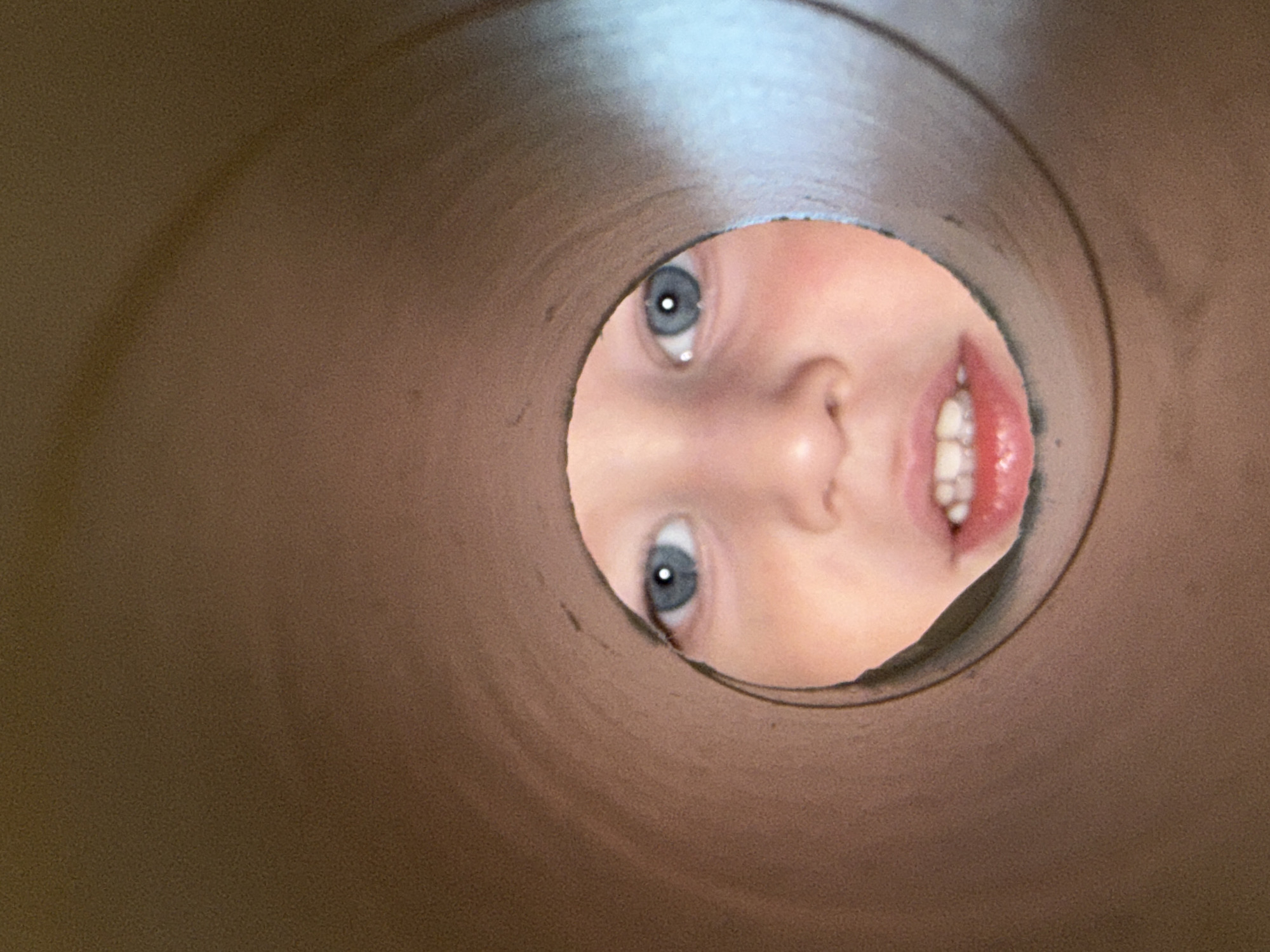 A close-up, slightly distorted photograph depicts a child's face peering through a circular opening. The child has fair skin, blue eyes, a small nose, and a partly open mouth revealing teeth and pink gums. The environment surrounding the child's face appears to be the interior of a brown, cylindrical tube or tunnel. The lighting is somewhat dim and the focus is mainly on the child's face. There is no evidence that reveals the location to be the city of Уральское, Russia.