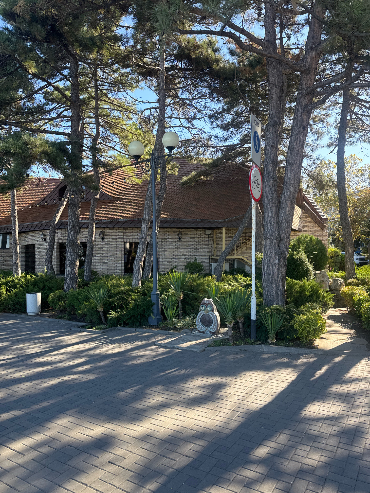 The image shows a view of a building with a tiled roof, constructed of light-colored bricks. The building is surrounded by numerous tall, mature trees with dense green foliage and visible pinecones. A paved walkway made of gray interlocking bricks runs in the foreground, with dappled sunlight and shadow patterns cast by the trees. To the left of the building, there is a white cylindrical trash can. In front of the building, a line of manicured green bushes and smaller palm-like plants is visible. A black lamppost with two globes stands near the center of the image, partially obscured by trees. On a white pole, there are two signs: a circular white sign with a blue silhouette of a person walking, and below it, a circular red and white sign with a bicycle symbol crossed out. To the left of the signs and bushes, a small mosaic sculpture resembling an owl sits on the ground. A narrow stone path leads away from the building to the right, disappearing into more greenery. In the background, a white car is partially visible. The sky is clear and blue. This scene is located in Anapa, Russia, likely in a park or a public area near a building.