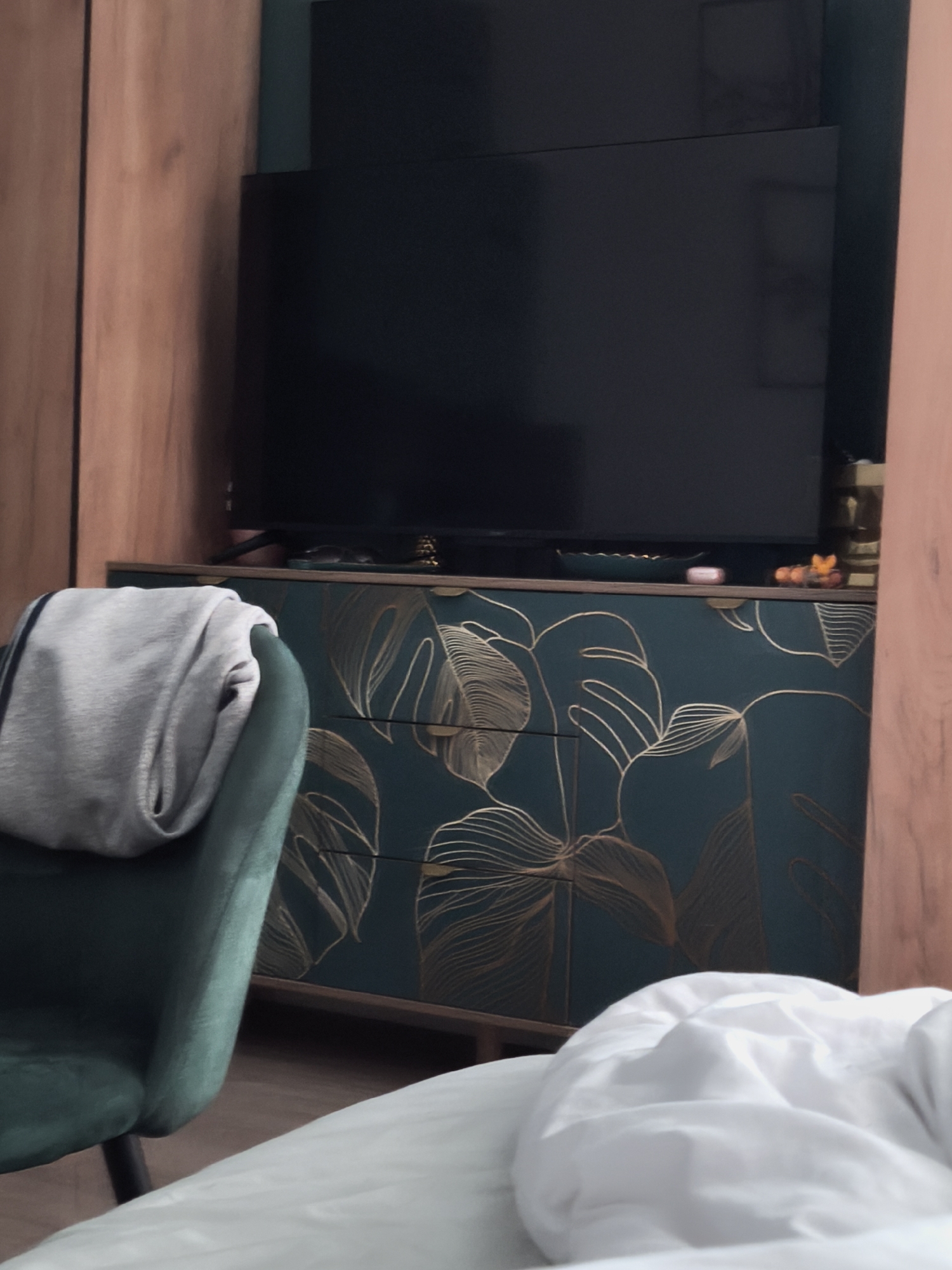 A dark screen television sits atop a dark green cabinet with gold monstera leaf detailing, in what appears to be a room in Moscow, Russia. A gray garment is draped over a green velvet chair. The lower right corner features rumpled white bedding. No people are visible.