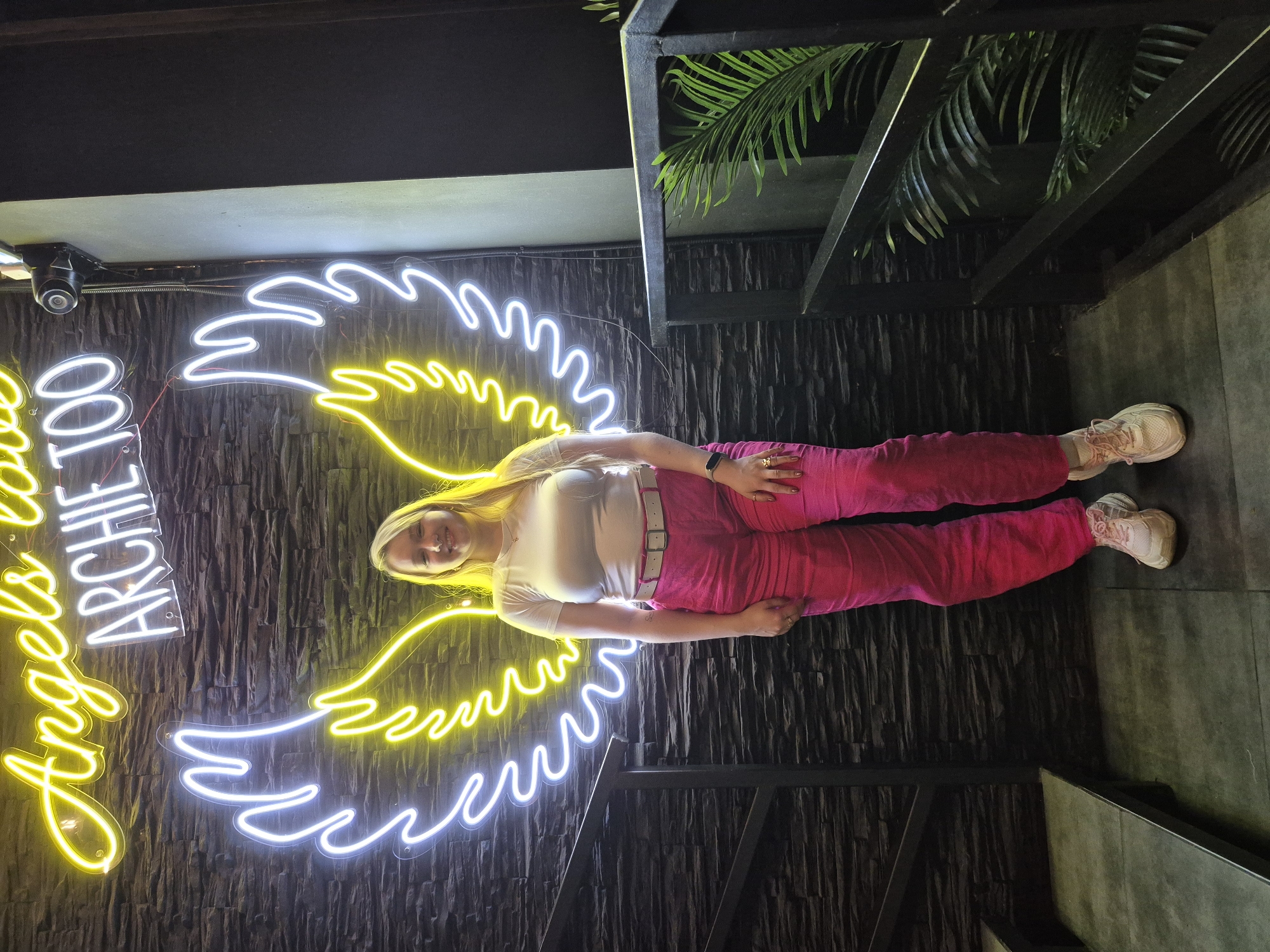 A young Caucasian woman with blonde hair, wearing a white t-shirt and bright pink pants, stands facing forward in front of a dark stone wall. Above her, a neon sign spells out "Angels love Archie TOO" in yellow and white light. To the left of the woman, white and yellow neon lights form a pair of angel wings, framing her. She is wearing a white belt with a gold buckle, and her shoes are light pink sneakers. The environment appears to be indoors, with a dark staircase and railing visible to the left and right of the woman. A potted palm plant is visible on the right side of the image. The woman is smiling.