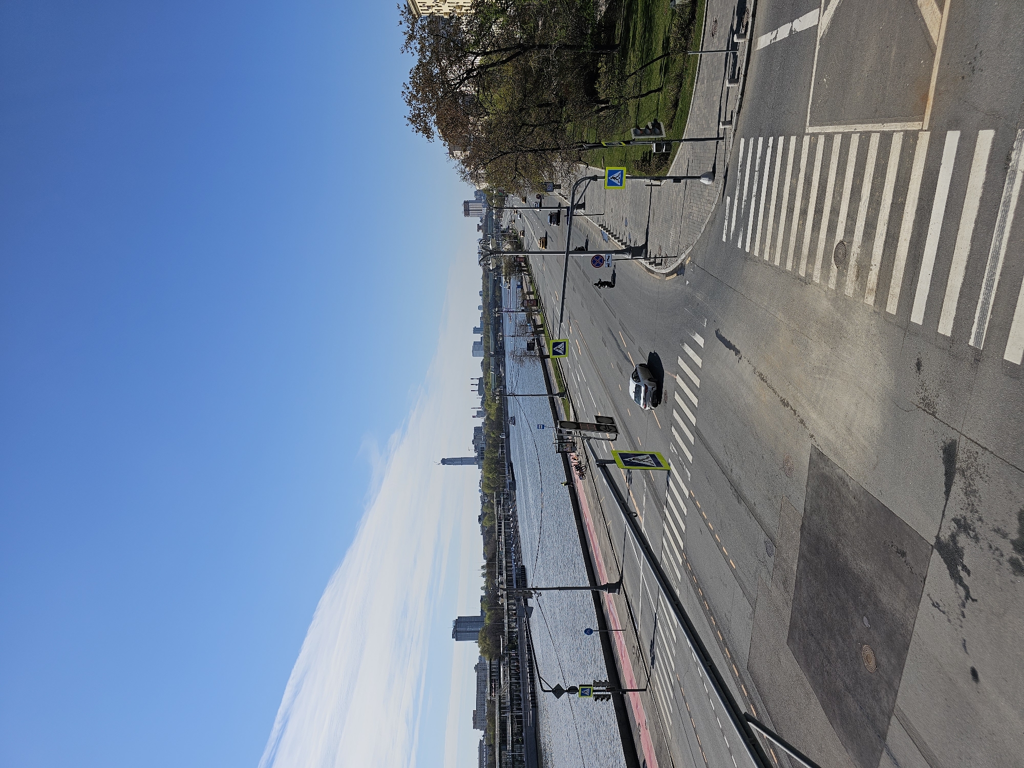The image shows a view of the Moskva River in Moscow, Russia. It is a sunny day, with a clear blue sky. The river is on the left side of the image, and the embankment and road are on the right. There is a white car driving on the road. Pedestrian crossings are visible on the road. There are streetlights and traffic signs along the embankment. Buildings can be seen in the distance across the river. There is greenery on the embankment, including trees and grass.