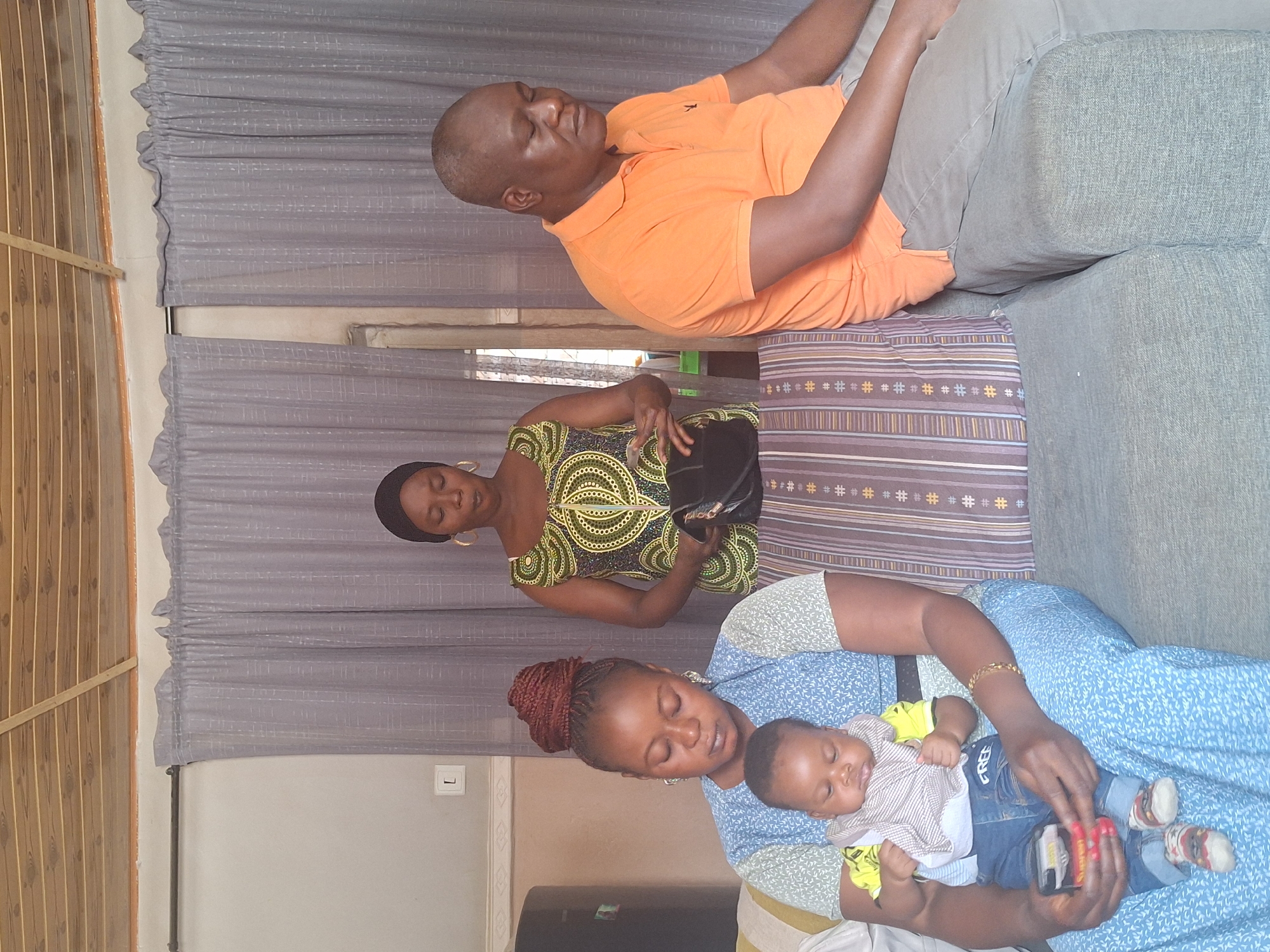 The image depicts three adults and an infant in an indoor setting. A man is seated on a grey sofa. Next to him, a woman in a patterned dress stands. In the foreground, a woman in a blue dress cradles an infant dressed in jeans and a striped top. The background includes a grey curtain and wooden framework, suggesting a window or room divider. The scene is likely situated in a domestic interior in Sabon Tasha, Nigeria, based on the cultural context reflected in the subjects and the attire.