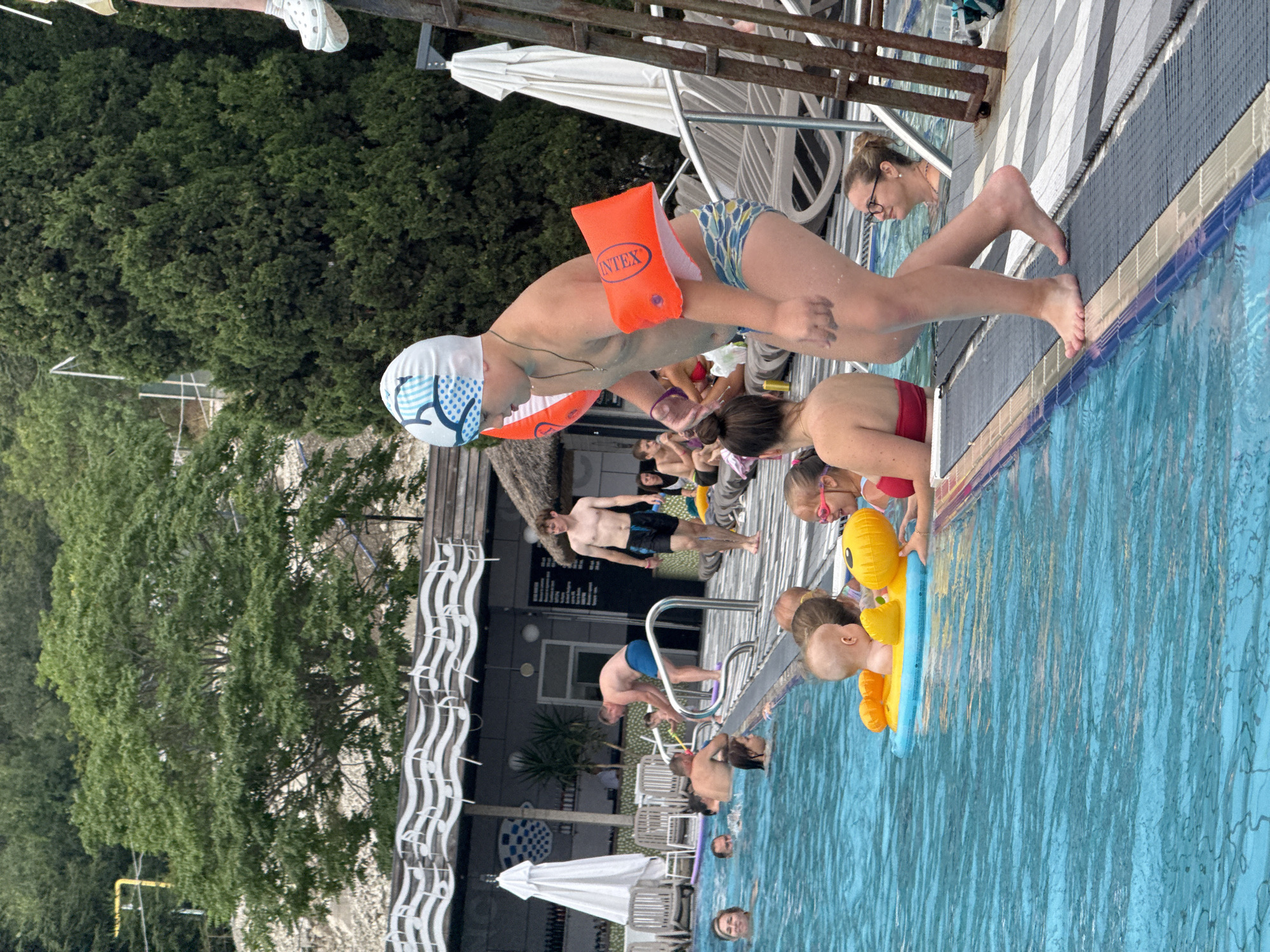 The image depicts a lively scene at an outdoor swimming pool in Yalta, Ukraine. In the foreground, a young boy wearing a swim cap and orange arm floaties stands on the edge of the pool, appearing ready to enter. He is wearing patterned swim trunks. Several other people are visible in and around the pool. A baby is floating in a yellow inflatable ring, with a young girl in a red swimsuit next to it. In the background, various adults and children are enjoying the water and relaxing. The pool area is surrounded by lounge chairs, umbrellas, and lush greenery, suggesting a resort or recreational facility. The architecture of the buildings in the background features decorative musical notes on a canopy, adding a unique touch to the environment.