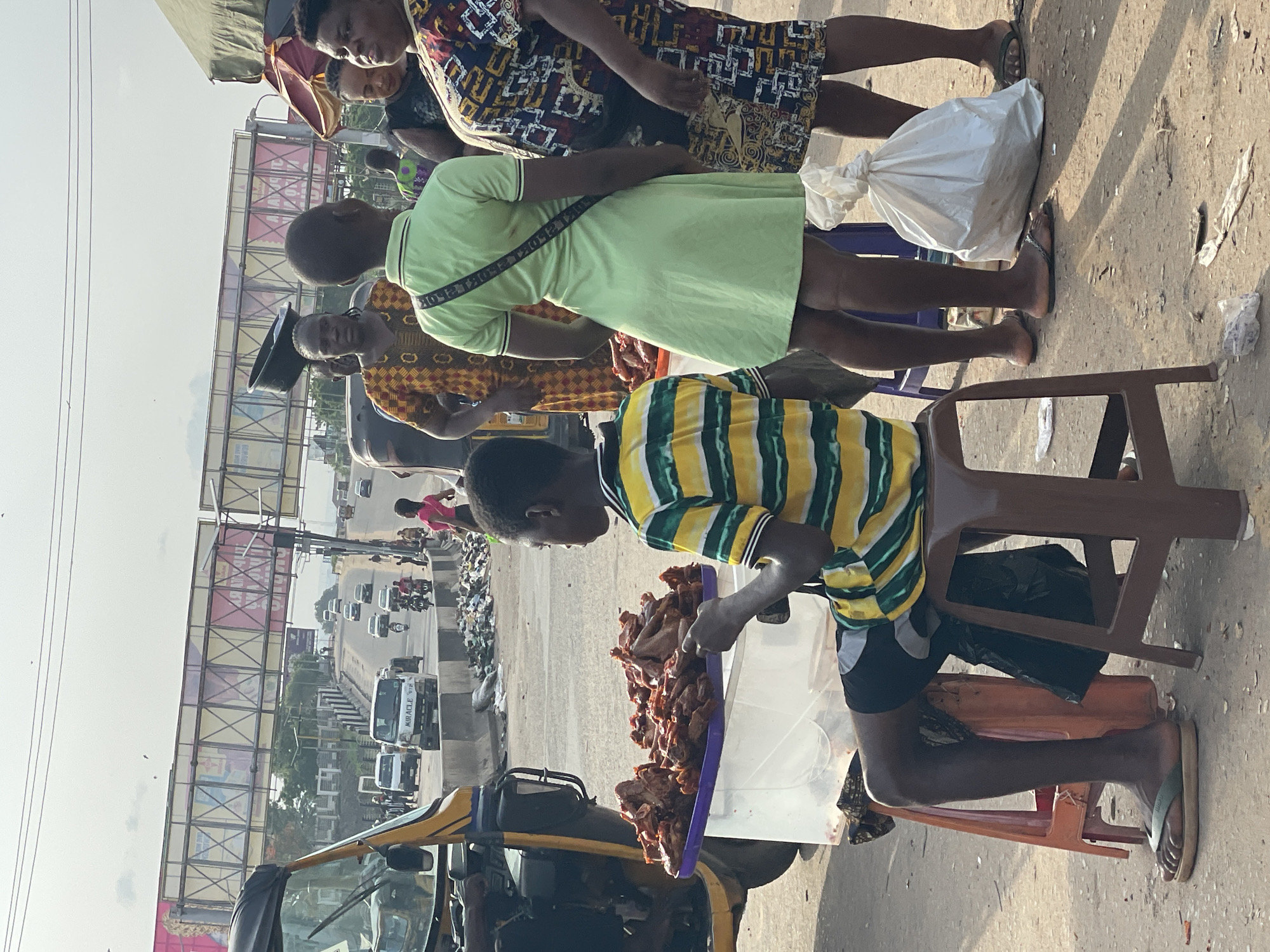 The image shows a street scene in Nkpor, Nigeria. A young man wearing a striped yellow and green shirt sits on a plastic chair next to a tray of what appears to be cooked meat. Several people stand nearby, presumably customers. A yellow auto rickshaw is parked next to the vendor. In the background, a bridge spans a road with traffic, with billboards visible above. The area appears to be a busy market or roadside vending location.