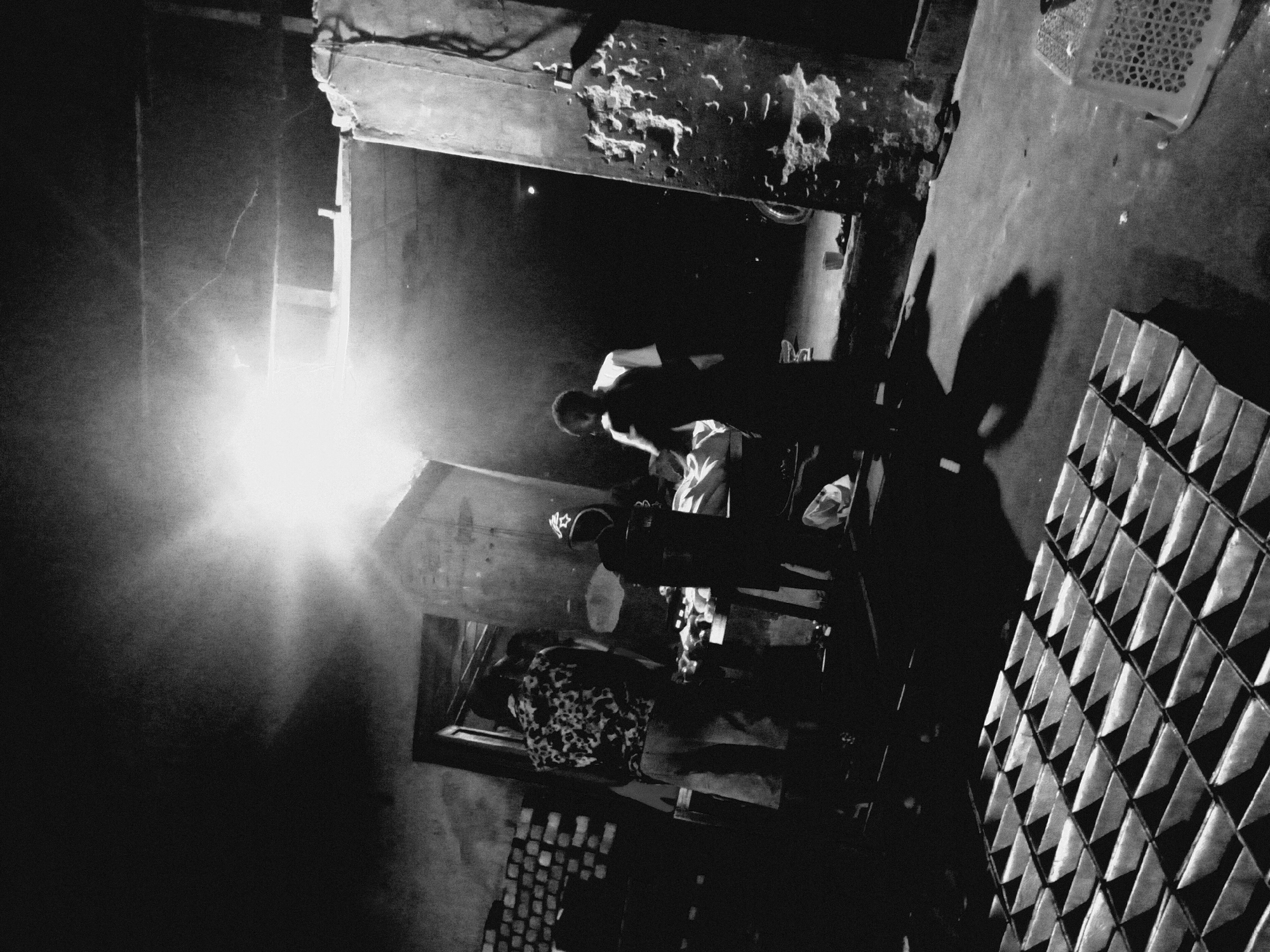 A high-contrast, grainy black and white photograph captures a dimly lit scene, likely at night or indoors, with several individuals engaged in activity. The primary light source, an intensely bright lamp or bulb, creates significant lens flare on the left, casting stark shadows and illuminating dust or smoke particles in the air.
In the foreground and center, several people are visible. One individual is hunched over, strongly backlit by the main light source, suggesting focused work or inspection. Another person stands further back, also partially silhouetted. Details of their faces are obscured by the lighting, but their postures convey engagement. One person wears a distinctively patterned shirt.
The setting appears to be a utilitarian or industrial space, characterized by rough textures on walls and an overhead beam showing signs of wear and crumbling material. To the right, a distinctive pattern of repeating triangular shapes forms a grid, possibly stacked objects like molds, bricks, or product packaging. Another woven or gridded container, resembling a crate, is also visible. The floor is a solid, relatively plain surface, marked by the strong, elongated shadows of people and unseen objects. No visible text is present in the image. The overall mood is dramatic and industrious, emphasizing form and contrast through its monochrome palette.
