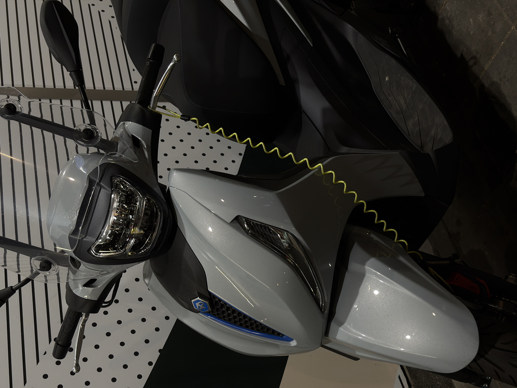 An upright, medium shot captures a close-up of the front and right side of a light gray scooter, parked indoors. The scooter features a large windshield, a prominent headlight assembly with intricate detailing, a silver brake lever, and a black handlebar grip. A bright yellow coiled security cable is attached to the handlebar and extends downwards towards the seat. The scooter's front fender, also in light gray with a metallic sheen, is visible. The background consists of a wall with a white and black polka dot pattern on the left, and a black surface with horizontal white stripes on the top. The floor appears to be tiled with dark gray rectangular tiles. The scene is dimly lit, with some highlights reflecting off the scooter's metallic surfaces. The scooter is positioned with its front facing slightly left. There are no people or animals visible in the image. The environment suggests an indoor setting, possibly a showroom or exhibition space. The scooter is a Piaggio model, indicated by the logo on the front grille. The location is identified as Vracar Urban Municipality, Serbia, although no specific visual cues in the image directly confirm this location.