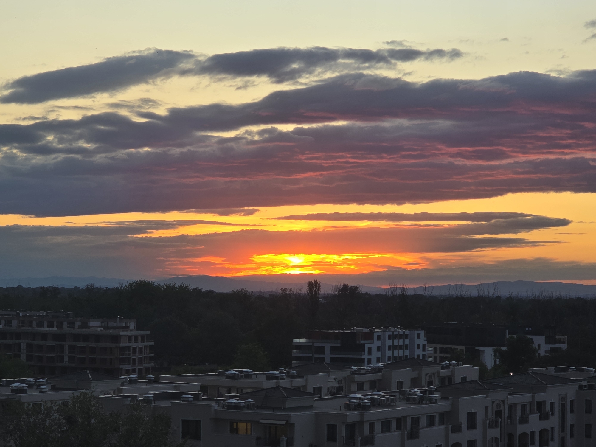 The image depicts a sunset over a cityscape. The sky is filled with clouds, exhibiting colors of yellow, orange, and pink near the horizon. The sun is partially obscured by the clouds, creating a vibrant glow. Below the horizon, there is a line of dark trees and a range of distant hills or mountains. The foreground is dominated by rooftops of buildings with white or light-colored walls and dark roofs. Some buildings are still under construction with exposed frameworks. The city name and location are not discernible.