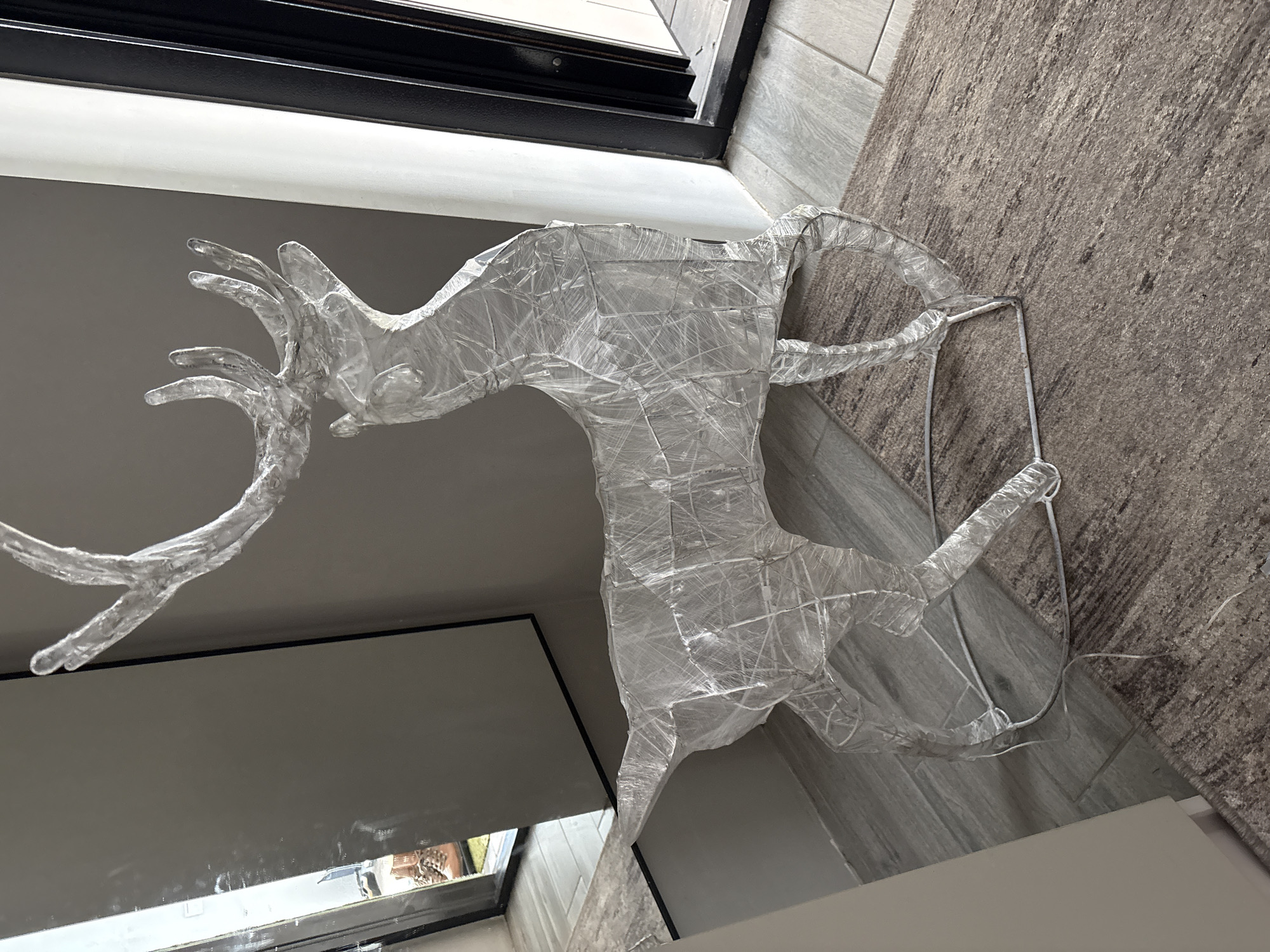 A translucent, approximately life-sized reindeer sculpture, constructed from a wire frame covered in crinkled clear plastic sheeting, stands on a textured brownish-grey area rug. The rug is positioned over light grey rectangular floor tiles. The sculpture faces left, its head slightly elevated, featuring prominent multi-branched antlers. The background consists of plain light grey walls. To the left, a vertical rectangular mirror with a black frame reflects a pair of brown laced boots and several pinecones, consistent with an entryway. To the right, a dark-framed doorway is partially open, revealing a lighter floor surface beyond. No individuals are present in the scene. The location is Votkinsk, Russia.