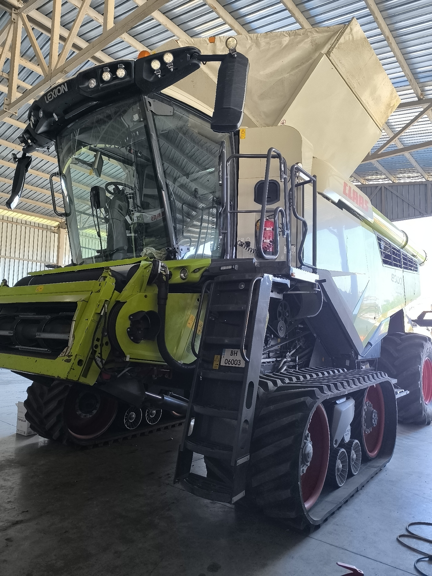 A Claas Lexion combine harvester, identified by the "LEXION" branding on its cabin and the "CLAAS" logo on its body, is parked indoors, likely in a maintenance shed or barn, in the village of Maior Uzunovo, Bulgaria. The combine is predominantly light green and beige, with black tracks on the left side and a large tire with red rim on the right. It features a large, transparent cabin with visible controls and seats inside. A ladder is attached to the side of the combine, leading up to the operator's area. A fire extinguisher is mounted on the side of the machine. The environment appears to be a utilitarian space with a corrugated metal roof supported by wooden beams and metal framing. The floor is concrete. The overall impression is of a large agricultural machine at rest in a functional setting.