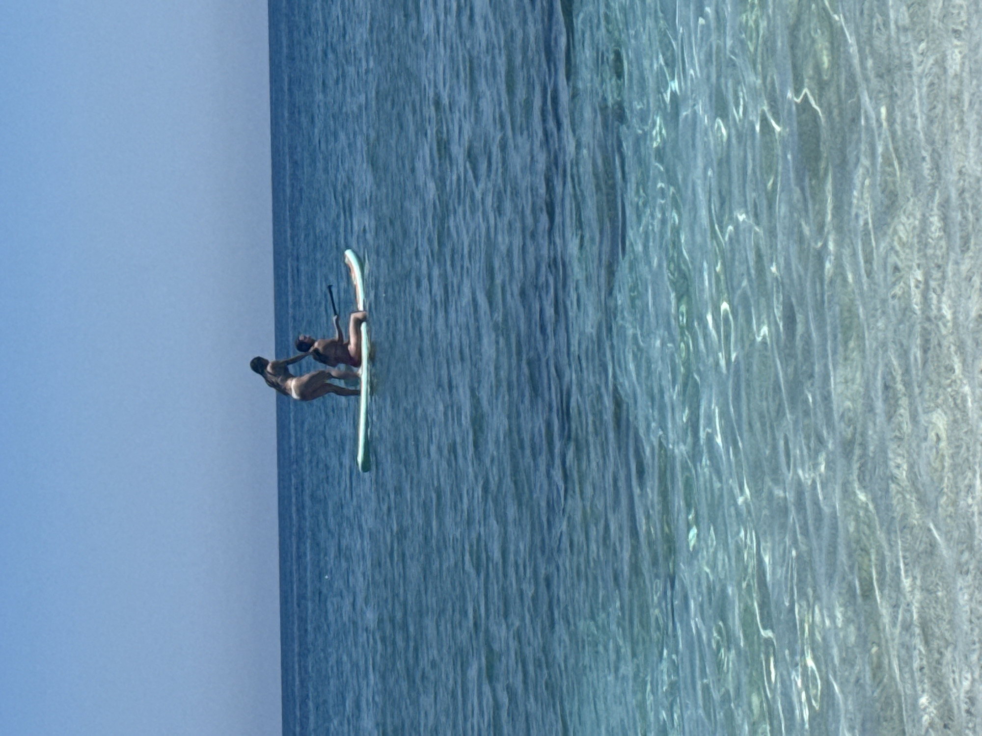 In the azure waters of Chersonisos Municipal Unit, Greece, two individuals engage in paddleboarding. One person, positioned towards the rear of the paddleboard, stands upright with arms outstretched. The second individual is seated on the paddleboard, holding a paddle, seemingly preparing to propel the board forward. The crystal-clear water reveals the sandy seabed beneath, with gentle ripples disturbing the otherwise tranquil surface. The sky above is a clear, cloudless blue, denoting a bright, sunny day.