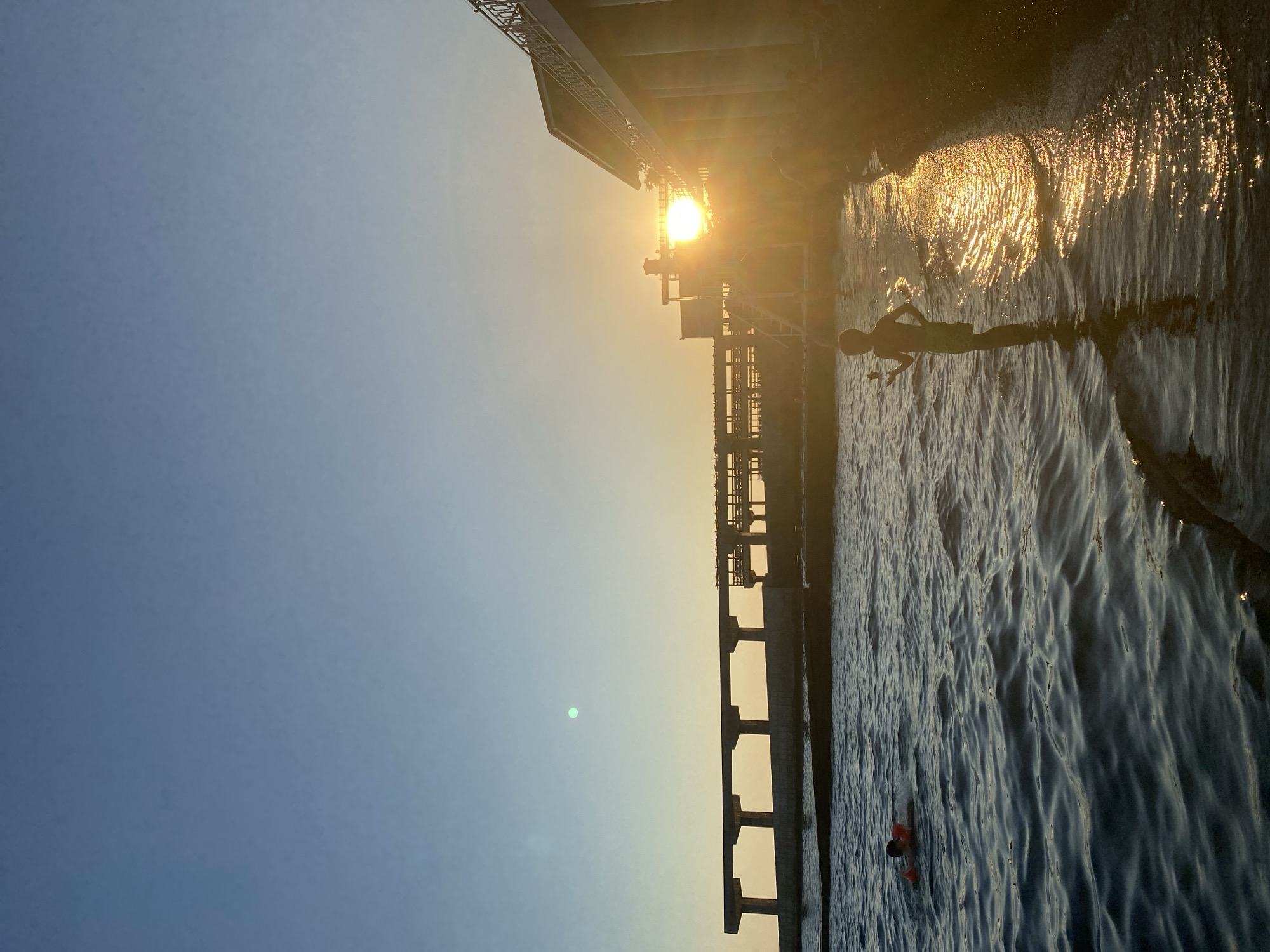 The image depicts a scene at a seaside location, likely Sochi, Russia, during sunset. The sun, low in the sky and partially obscured by an overhead structure, casts a golden glow across the water and the sky. The water's surface is rippled, with sunlight creating a shimmering effect.
In the foreground, a silhouette of a person is visible in the water, appearing to be swimming or wading. Further in the distance, another person can be seen in the water, wearing what appears to be a life vest.
An overhead structure, possibly a pier, bridge, or part of a building, dominates the upper portion of the image, with its supports and railings casting shadows. The sky transitions from a clear blue at the top to a warm yellow-orange hue near the horizon, indicating the time of day is late afternoon or early evening. The overall mood of the image is serene and tranquil, with the warm sunlight creating a peaceful atmosphere. No visible text is present in the image.