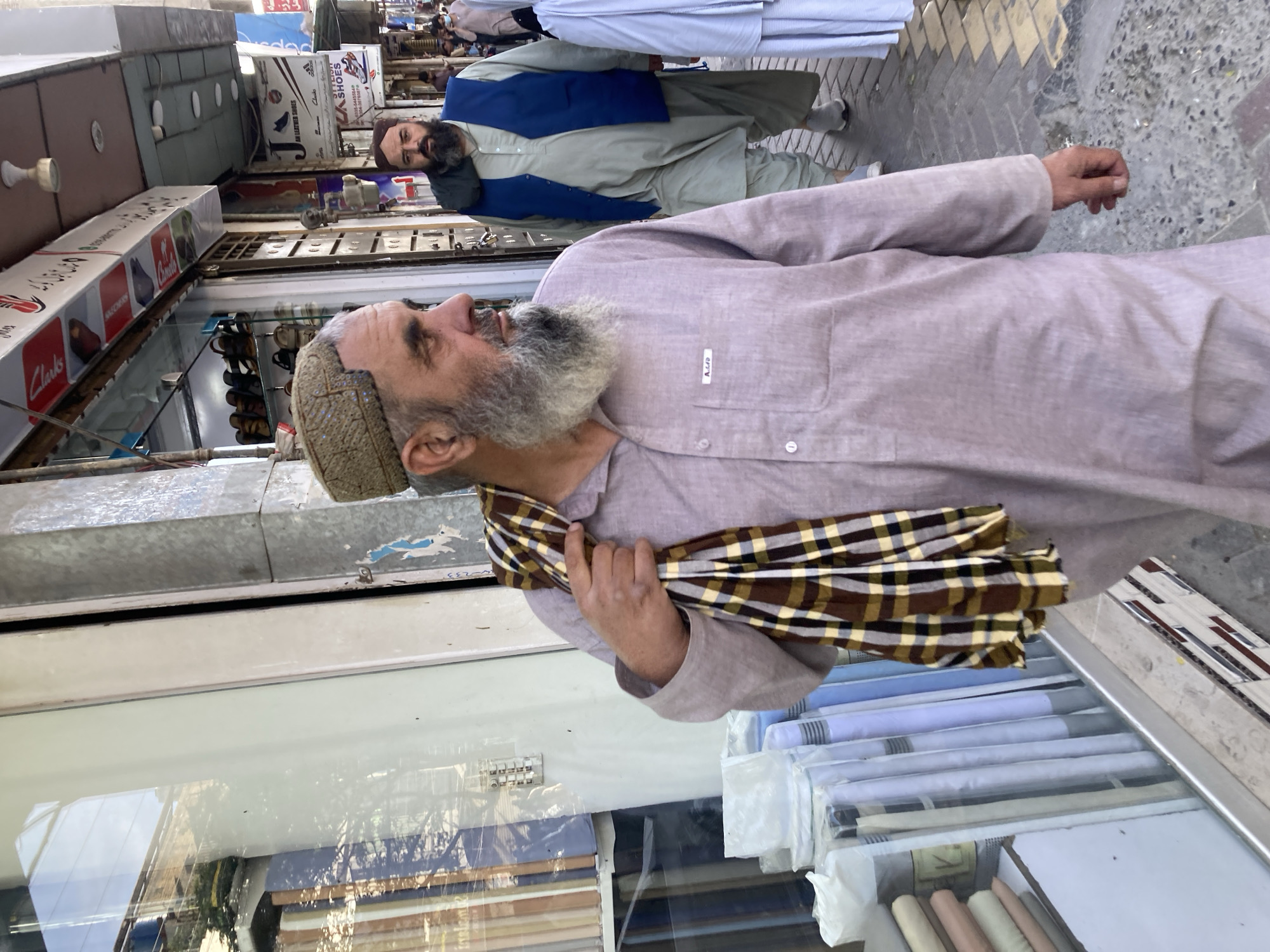 A medium, eye-level shot depicts a man in Quetta City Tehsil, Pakistan. The man, who appears to be of middle-eastern or central-Asian descent, has gray hair and a gray beard. He wears a gray-brown hat and a long-sleeved gray-brown shirt with a plaid scarf wrapped around his upper chest. The man is looking to his left with his right hand extended outwards. Behind him, another man with a beard is wearing a blue vest over a light-gray outfit. They are on a sidewalk alongside a store. The store displays shoes and fabric. The scene suggests an urban environment, typical of a commercial street.