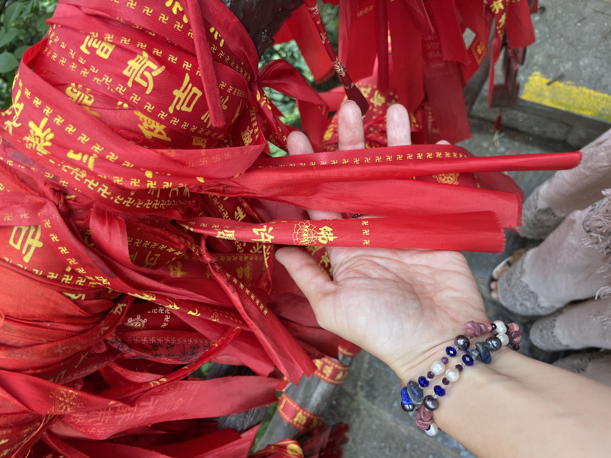 A close-up captures a human hand amidst a cascade of vibrant red ribbons tied to a wooden structure, possibly a tree or post. The ribbons are adorned with yellow Buddhist swastikas and Chinese characters, suggesting a religious or cultural significance. The hand, which is partially open, holds one of the ribbons, allowing the fingers to be visible against the backdrop of cascading fabric. A beaded bracelet, featuring pearls, lapis lazuli, and other colorful stones, adorns the wrist of the hand. In the periphery, a glimpse of a person's feet in sandals is visible. This scene appears to be set in Luoguta, China, indicating a location where such cultural or religious practices may be observed.