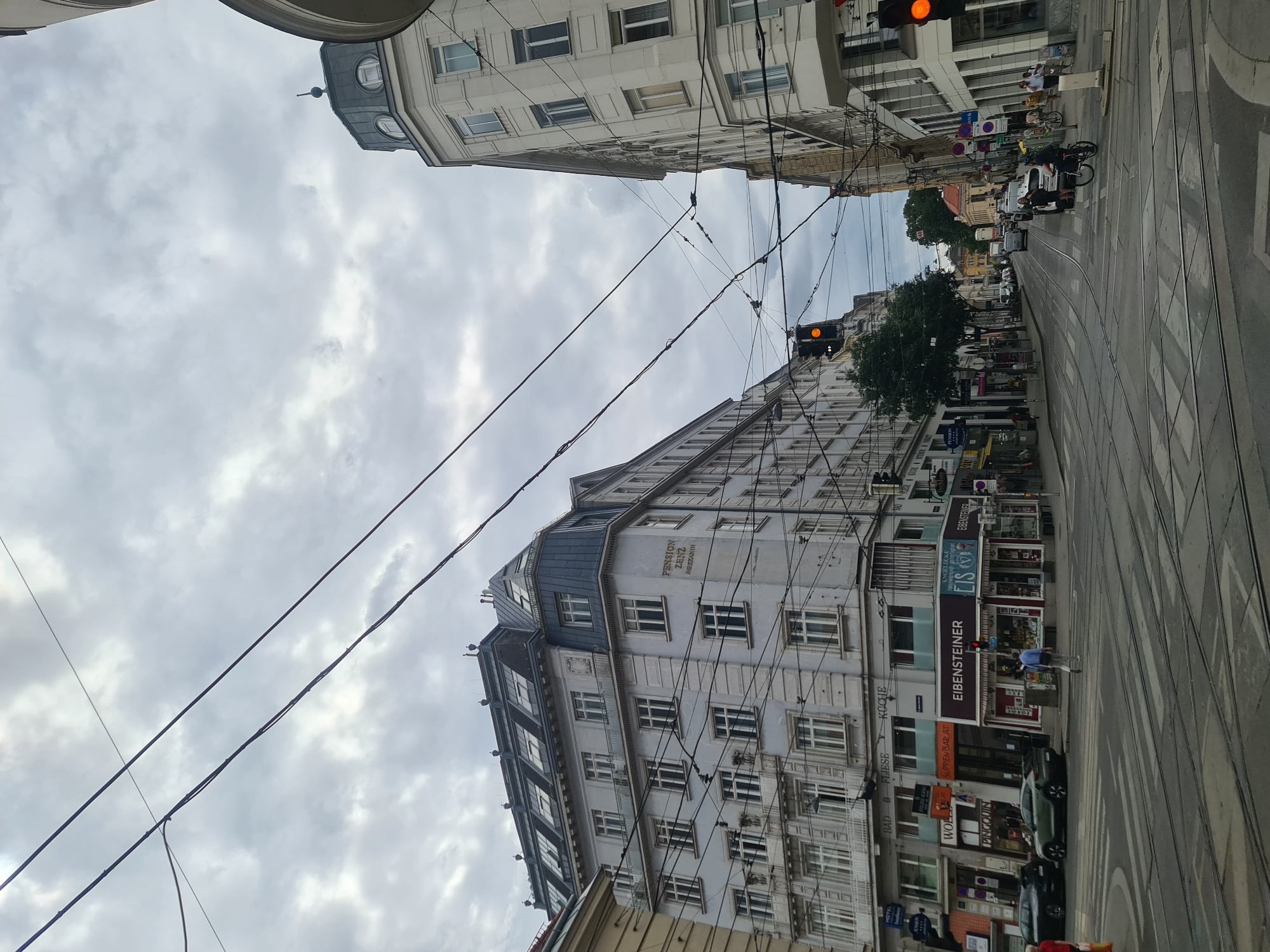 The image depicts a street scene in Vienna, Austria, under a cloudy sky. Overhead, a complex network of trolley wires crisscrosses the frame, indicative of the city's public transportation system. On the left, a multi-story building with a mansard roof and numerous windows displays signage for "PENSION ZENZ MEZZANIN." Below this, storefronts are visible, including one labeled "EIBENSTEINER" and another featuring the word "EIS."
In the foreground, a wide intersection with multiple tram tracks dominates the street. Pedestrian crossings are clearly marked. Several cars are present on the road, including a black sedan and a green SUV parked near the curb. A lone pedestrian walks across the intersection.
On the right side of the street, another multi-story building with rounded corner architecture stands. Traffic lights are visible, with one displaying an amber light. Further down the street, more vehicles, including cars and a bicycle, are moving through the intersection. The overall impression is a typical urban streetscape in a European city.