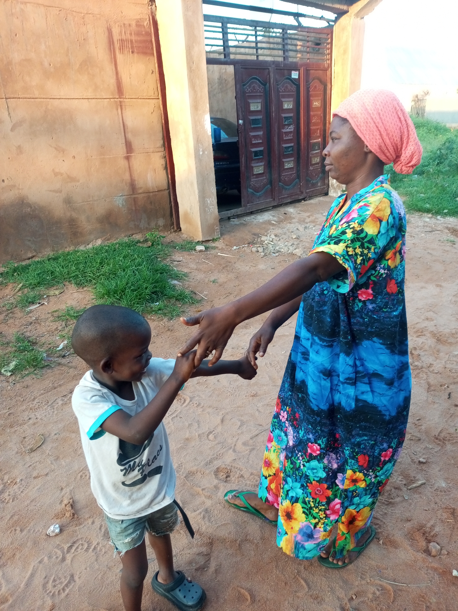 In the city of Tempure, Nigeria, a young boy in a light-colored t-shirt and denim shorts is holding hands with an adult woman who is wearing a brightly colored, floral patterned dress and a pink headscarf. They are standing on a dirt ground in front of a building with a concrete wall and a large, ornate double door. The building appears to be a residential structure. There is some green grass growing near the building. The scene is illuminated by natural light, suggesting it is daytime.