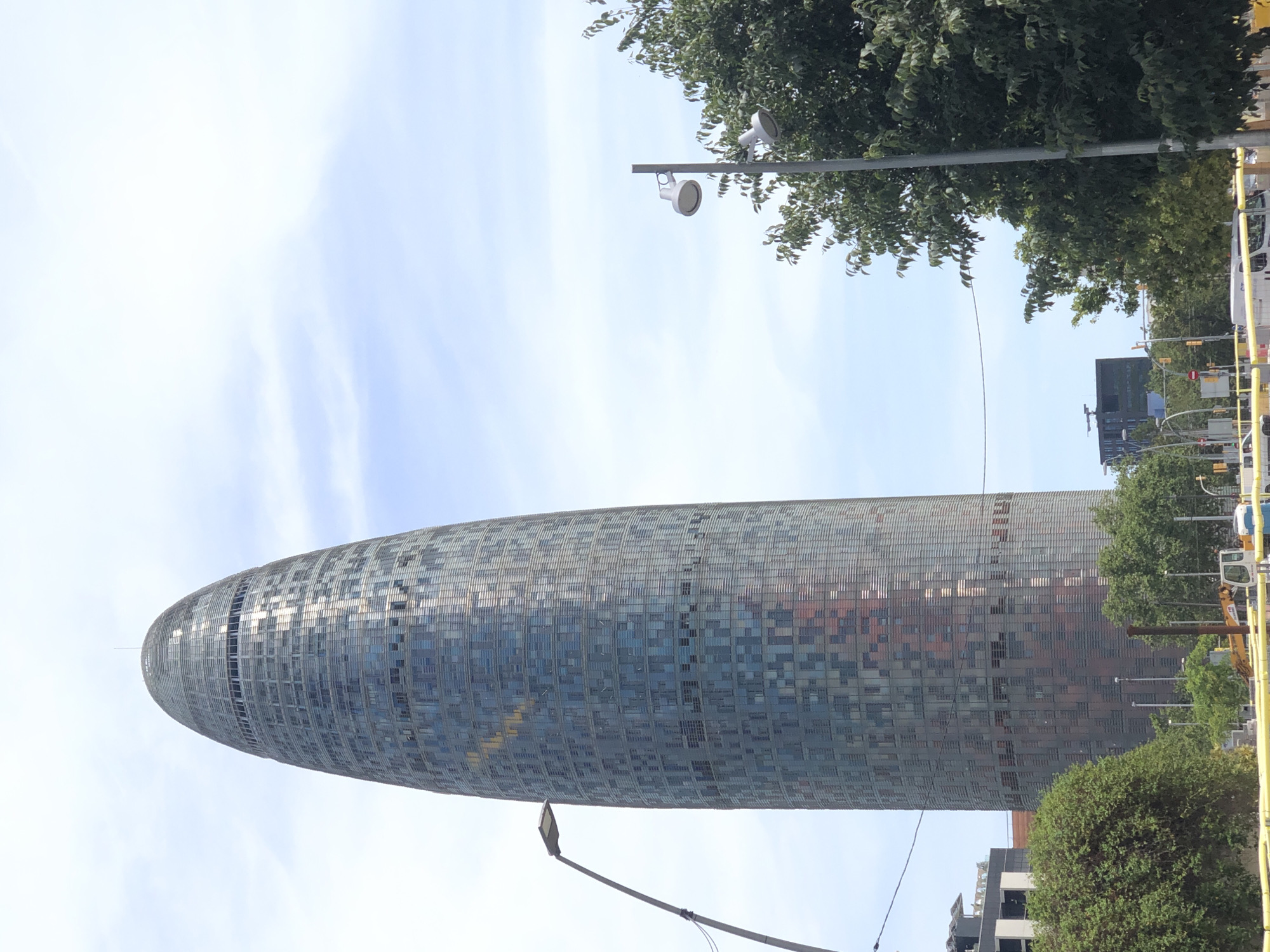 In an outdoor urban setting, a tall, cylindrical skyscraper with a rounded top dominates the frame. The building's facade is covered in a grid of windows, reflecting the sky and surrounding environment. A street lamp with a single light fixture stands to the left of the skyscraper, and another street lamp with two fixtures is visible in the middle ground to the right. Lush green trees are scattered throughout the scene, with dense foliage on the left and sparser trees on the right. The sky above is a pale blue with scattered white clouds. In the foreground, yellow barriers line what appears to be a construction or roadwork area, with glimpses of vehicles and equipment. The overall impression is a bright, sunny day in a modern city.