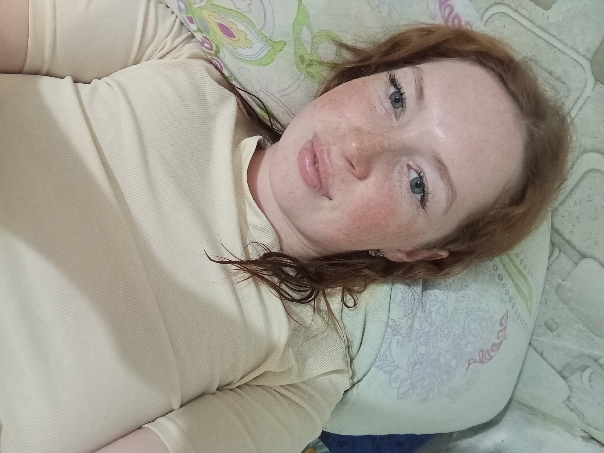 A fair-skinned woman with red hair and freckles is lying down, looking directly at the camera. She is wearing a light-colored top. She is on a pillow with a floral pattern, which in turn rests on a textured surface. The location is Yurgamysh, Russia.