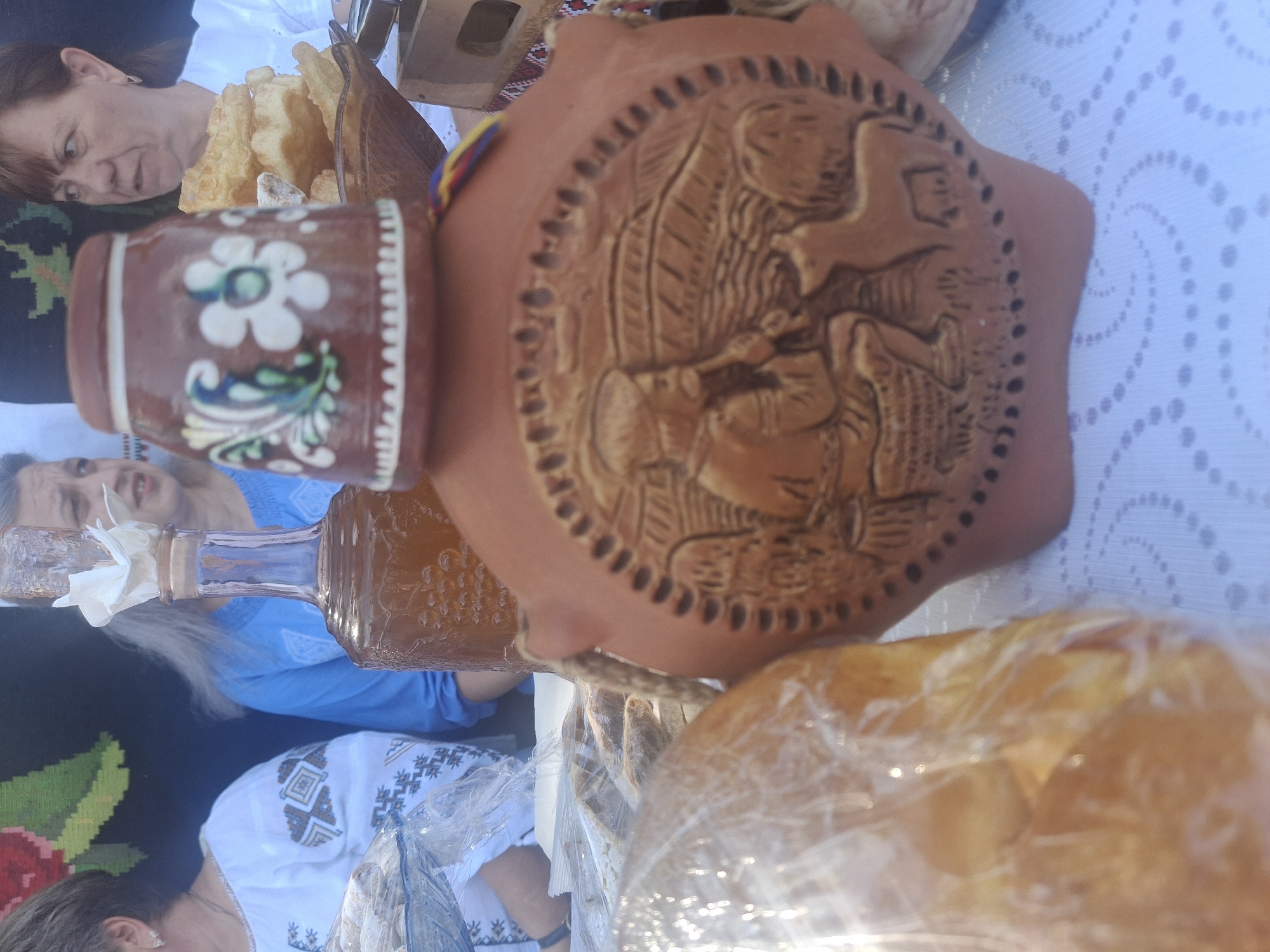 In Roghi, Moldova, an outdoor stall displays various items for sale. In the foreground, a large, terracotta flask with an embossed scene of a man playing a flute and a dog is prominently featured. Resting atop the flask is a smaller, decorated clay cup. To the left, a glass bowl contains what appears to be dried bread or pastries, wrapped in plastic. Further back, a glass bottle filled with amber liquid sits on a table. Three women are visible in the background. One, wearing a white embroidered shirt, is on the far left, partially obscured by the bowl. Another woman in a blue garment is in the center, and a third woman in a white shirt is on the right, looking towards the right edge of the frame. The stall is set up outdoors, suggested by the bright lighting and a woven tapestry with floral motifs in the background. A white tablecloth with a delicate spiral pattern covers the surface.