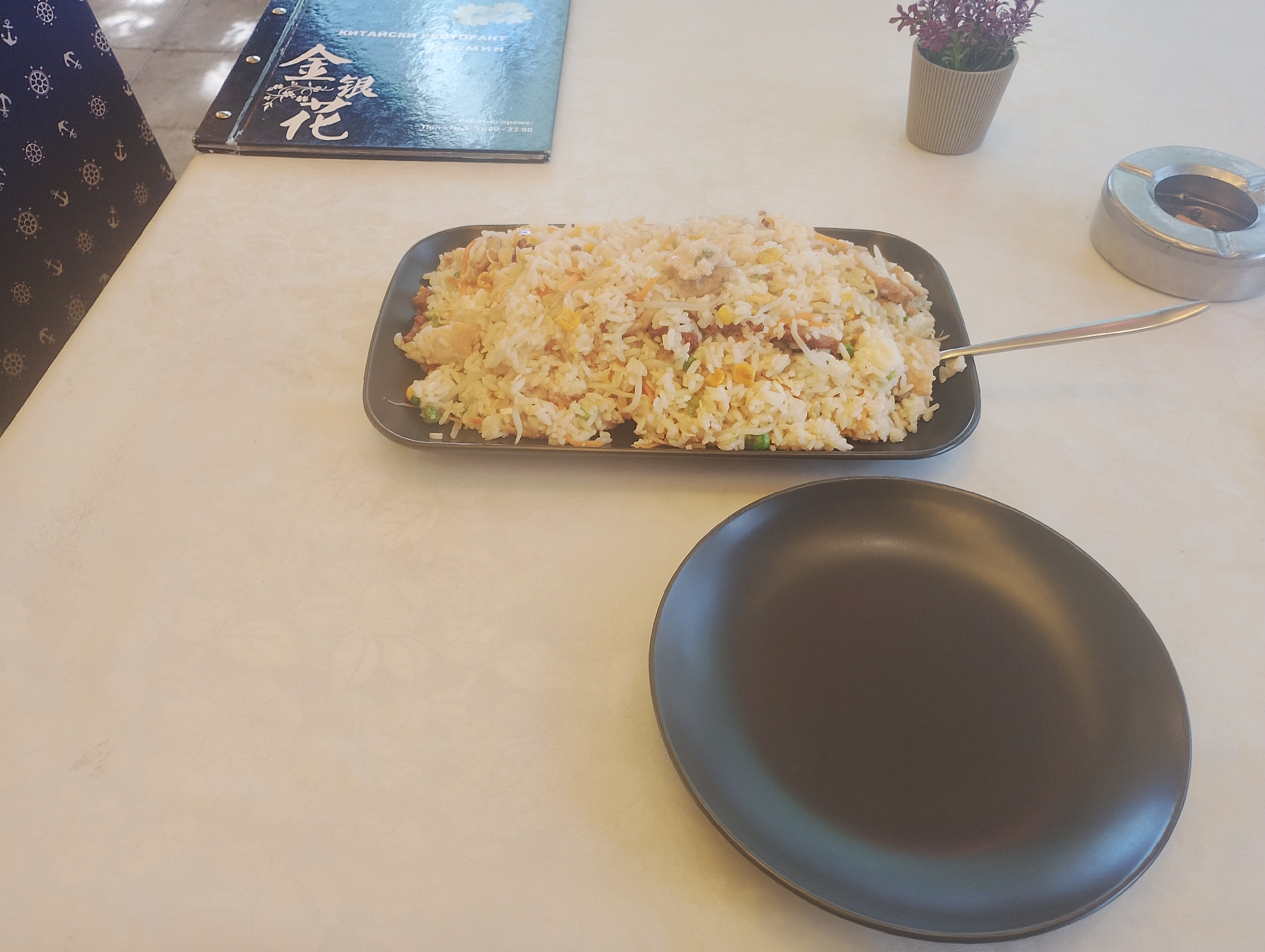 The image captures a dining table setting, likely at a restaurant. Dominating the scene is a large plate of fried rice, placed centrally on the table. A spoon rests on the edge of the plate, implying it's ready to be served. To the right of the rice, there's a small potted plant with purple flowers and a silver ashtray containing cigarette butts. A black plate is positioned below the fried rice, presumably for serving portions.
To the left, a partially visible menu with Chinese characters and Cyrillic text indicates it's a Chinese restaurant, specifically reading "Chinese Restaurant Golden Flower Restaurant" in Cyrillic. The restaurant's working hours are also shown. A portion of a navy blue chair with a white anchor and helm pattern is visible on the left side.
Given the Cyrillic script on the menu, it's plausible the restaurant is located in a country that uses this alphabet. Taking into account that the restaurant name is "Chinese Restaurant Golden Flower", and combining the information about the text of the menu, the picture was most likely taken in Burgas, Bulgaria.