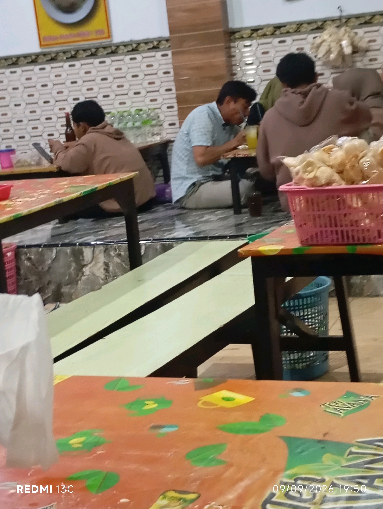 The image depicts an indoor scene, possibly a restaurant or food stall, in Wuluhan, Indonesia. In the foreground, a table with an orange, patterned surface is visible, with the bottom right corner displaying a timestamp "09/09/2026 19:50" and "REDMI 13C". Further back, another table with a similar patterned surface is set up. On this table, there are what appear to be fried snacks in a pink plastic basket. Behind this, a man in a light blue plaid shirt is seated at a table, holding a yellow cup or glass. Another person, wearing a brown hoodie, is seated next to him, facing away from the camera. A third person, also in a brown hoodie, is visible in the background, to the right. The environment features a patterned wallpaper on the wall and wooden furniture. The overall lighting suggests an indoor setting, possibly in the evening.