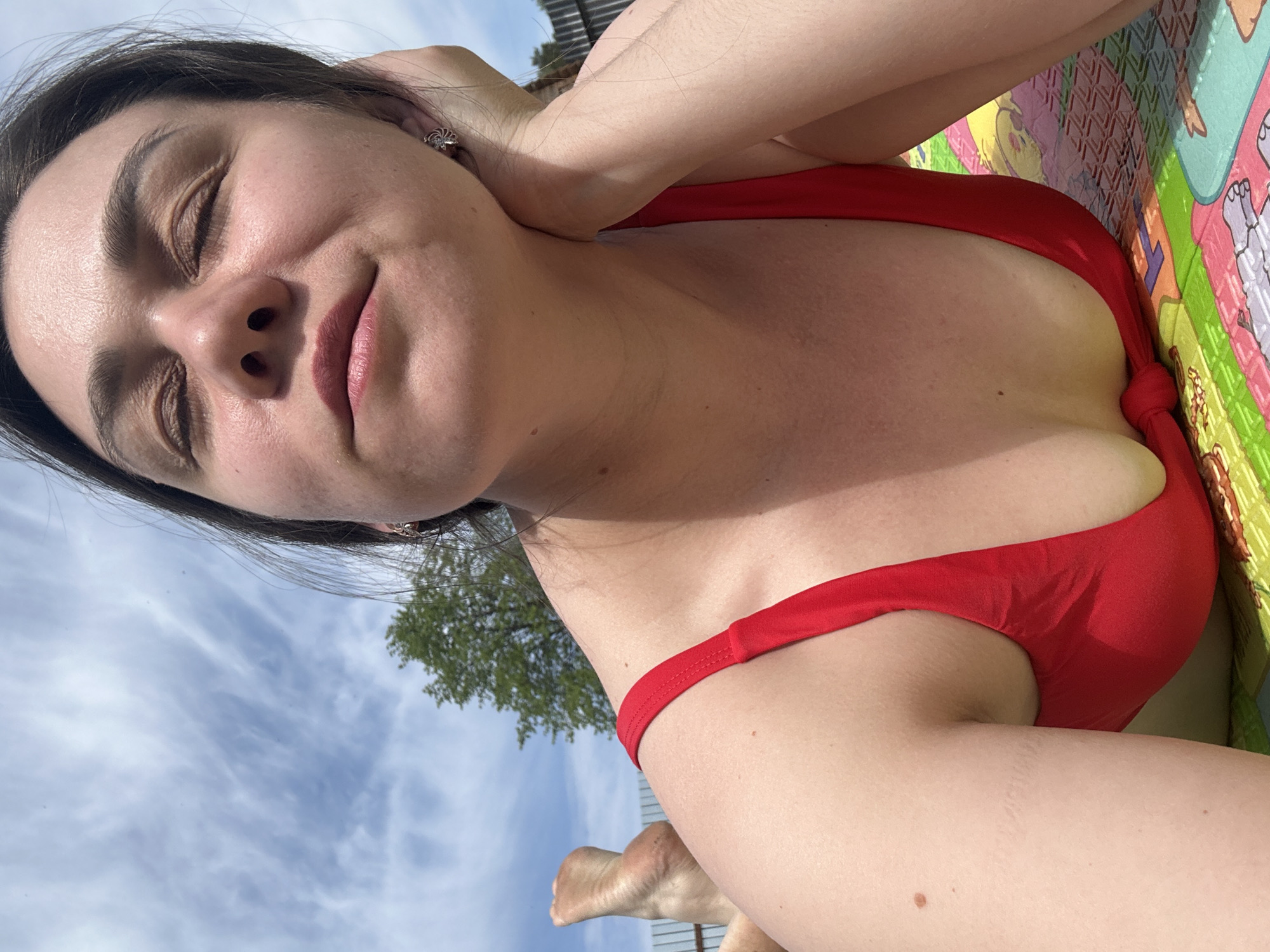 A woman with dark hair and fair skin is reclining outdoors in Chaikovsky, Russia. She has her eyes closed and appears relaxed. She is wearing a red swimsuit. She is on a colorful play mat. A tree and a cloudy sky are visible in the background.