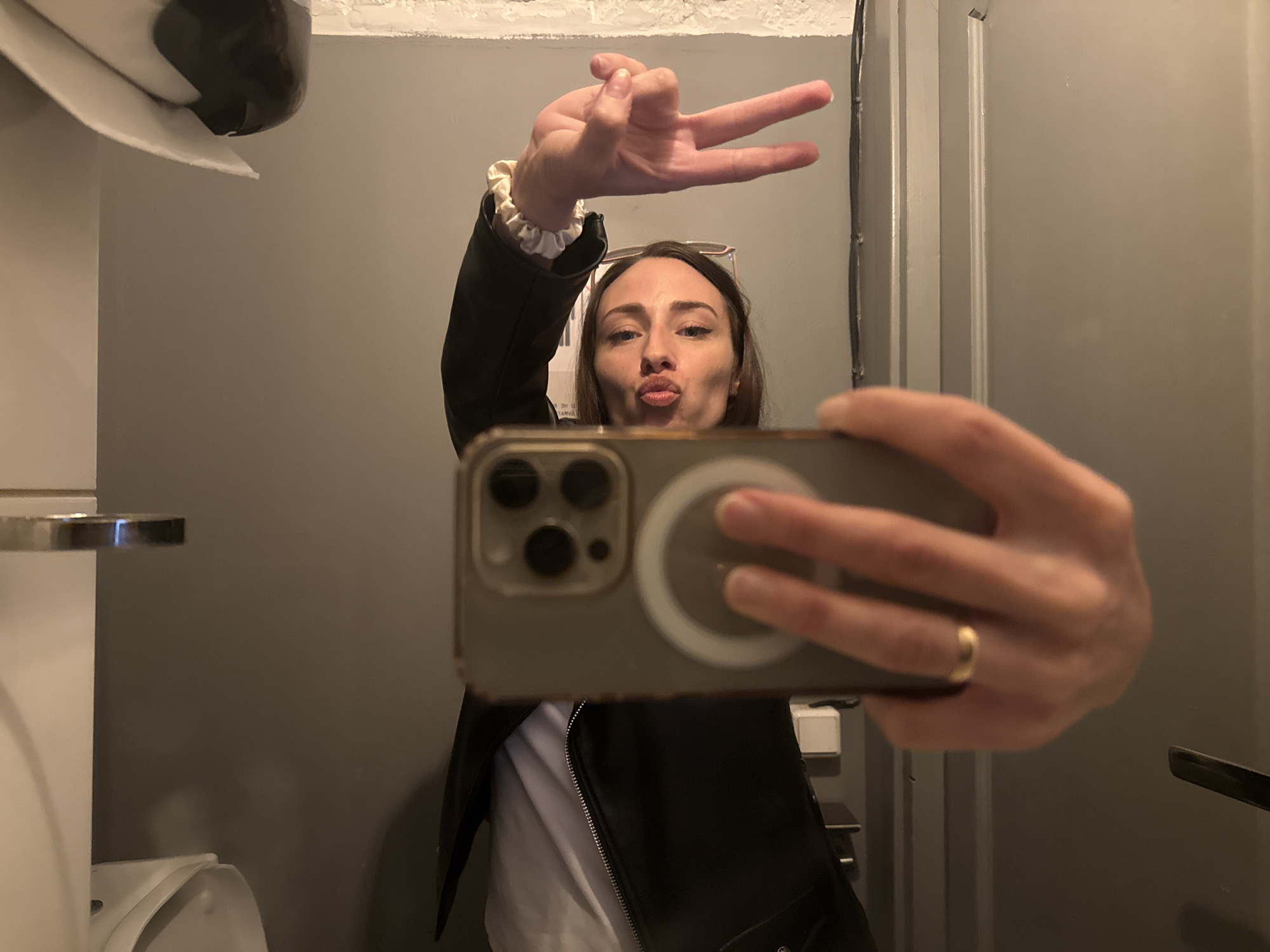 The image is a selfie taken in a bathroom. A person, presumably a woman, is holding up a smartphone, an iPhone, to capture the picture. She is puckering her lips in a kissing gesture and making a "peace" sign with her other hand. The subject is wearing a black leather jacket over a white top. There are glasses sitting on her forehead. On her finger she is wearing a gold ring. A toilet and toilet paper dispenser are visible in the periphery. The bathroom has gray walls and appears to be in a public or commercial setting. There is no indication of Saint Petersburg, Russia in the picture.