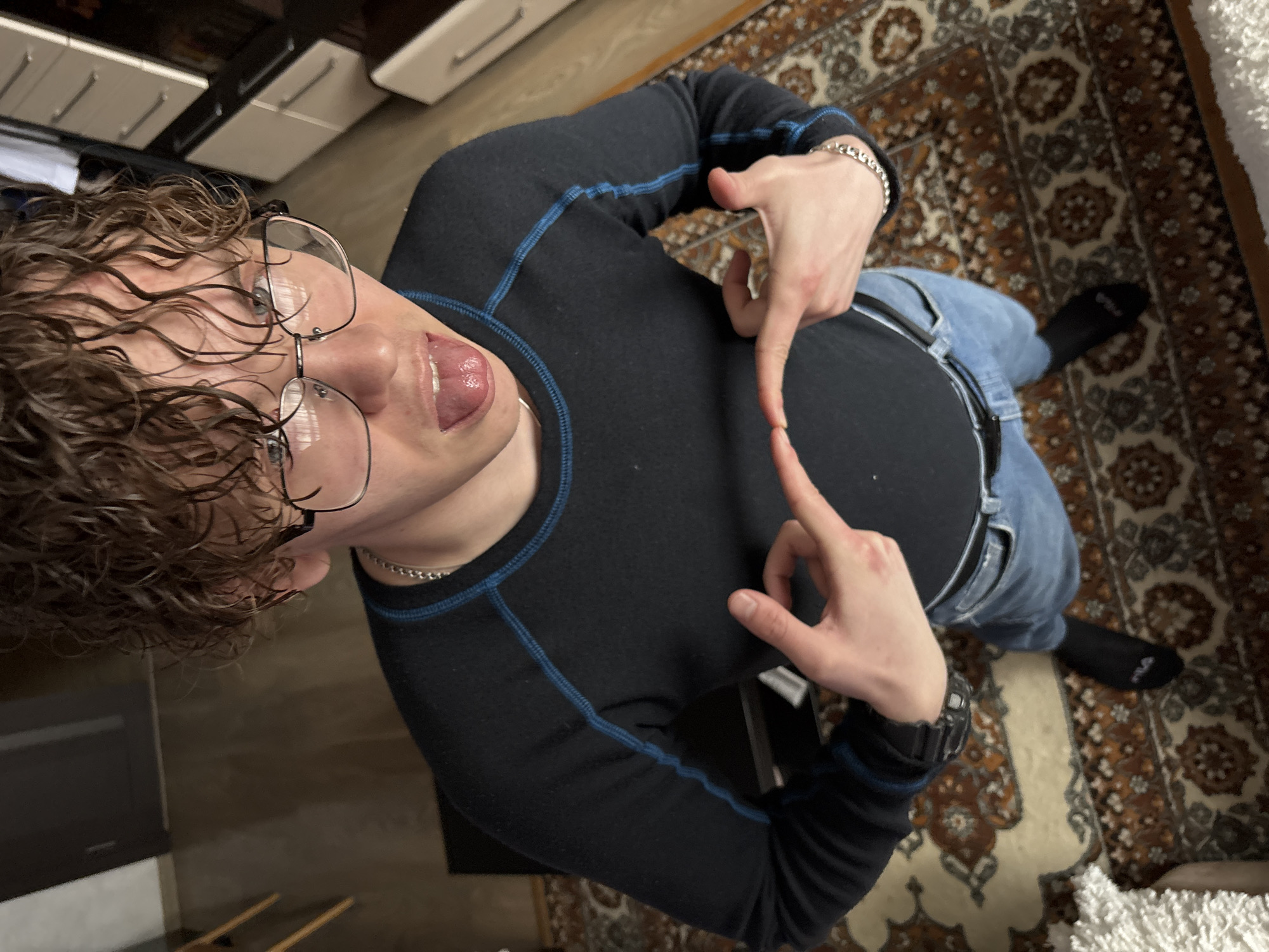 A person with light brown curly hair, wearing glasses, a black long-sleeved shirt with blue stitching, and light blue jeans, is sticking their tongue out and making a gesture with their hands, pointing at their chest. They are standing on a patterned rug with black socks. The floor appears to be made of dark wood. In the background, there are kitchen cabinets. The exact location in Perm, Russia, cannot be determined from the image alone.