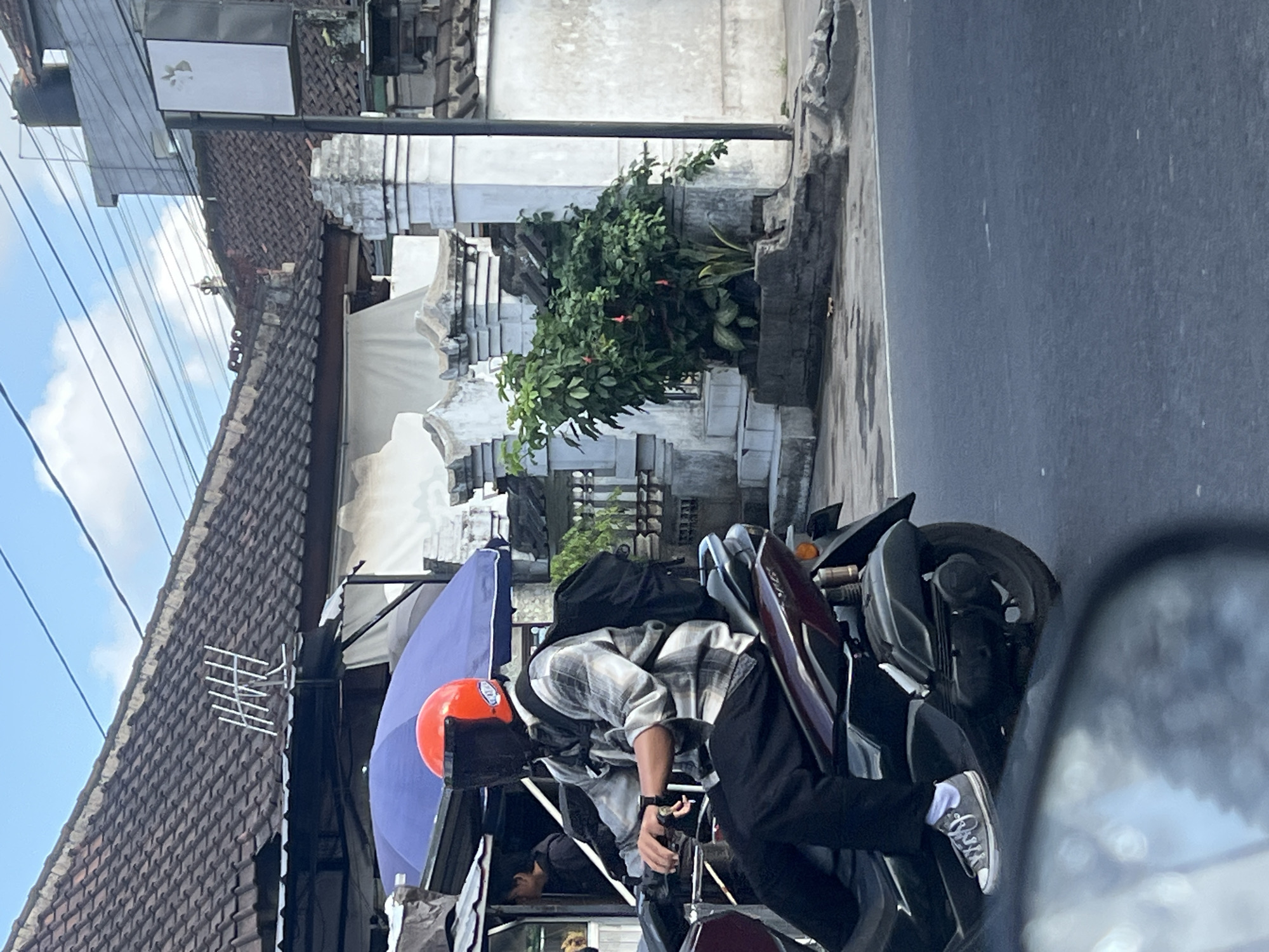 A man wearing an orange helmet with a black visor, a checkered shirt, and dark pants rides a dark-colored scooter on a street in Denpasar, Indonesia. He has a black backpack on. The scooter has a dark maroon body. In the background, there are buildings with tiled roofs, a blue umbrella, and lush green plants. The sky is partly cloudy.